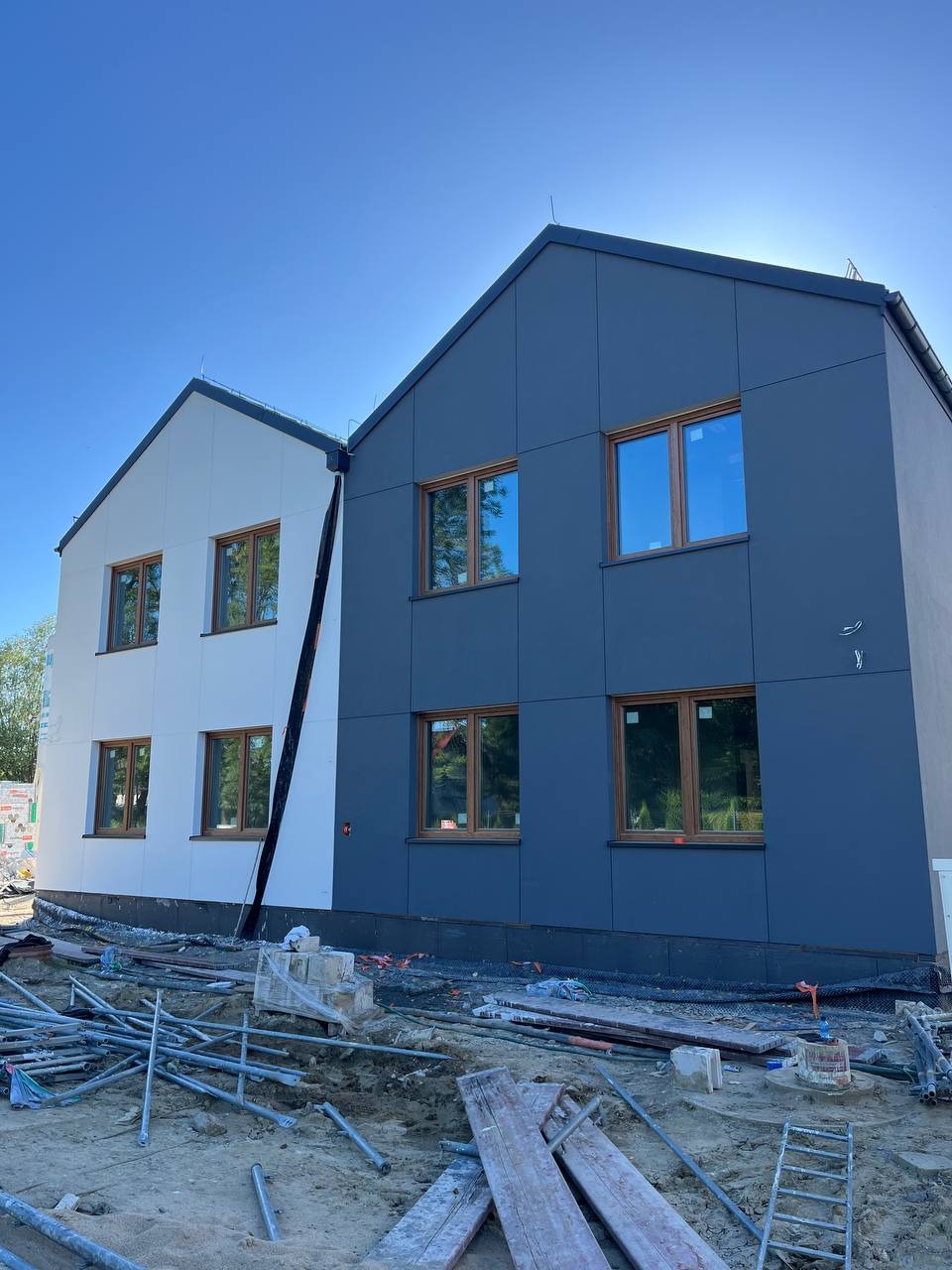 Dwa budynki w trakcie wykańczania elewacji: jeden biały, drugi grafitowy. Widoczne rusztowania i materiały budowlane. Okna z drewnianymi ramami.