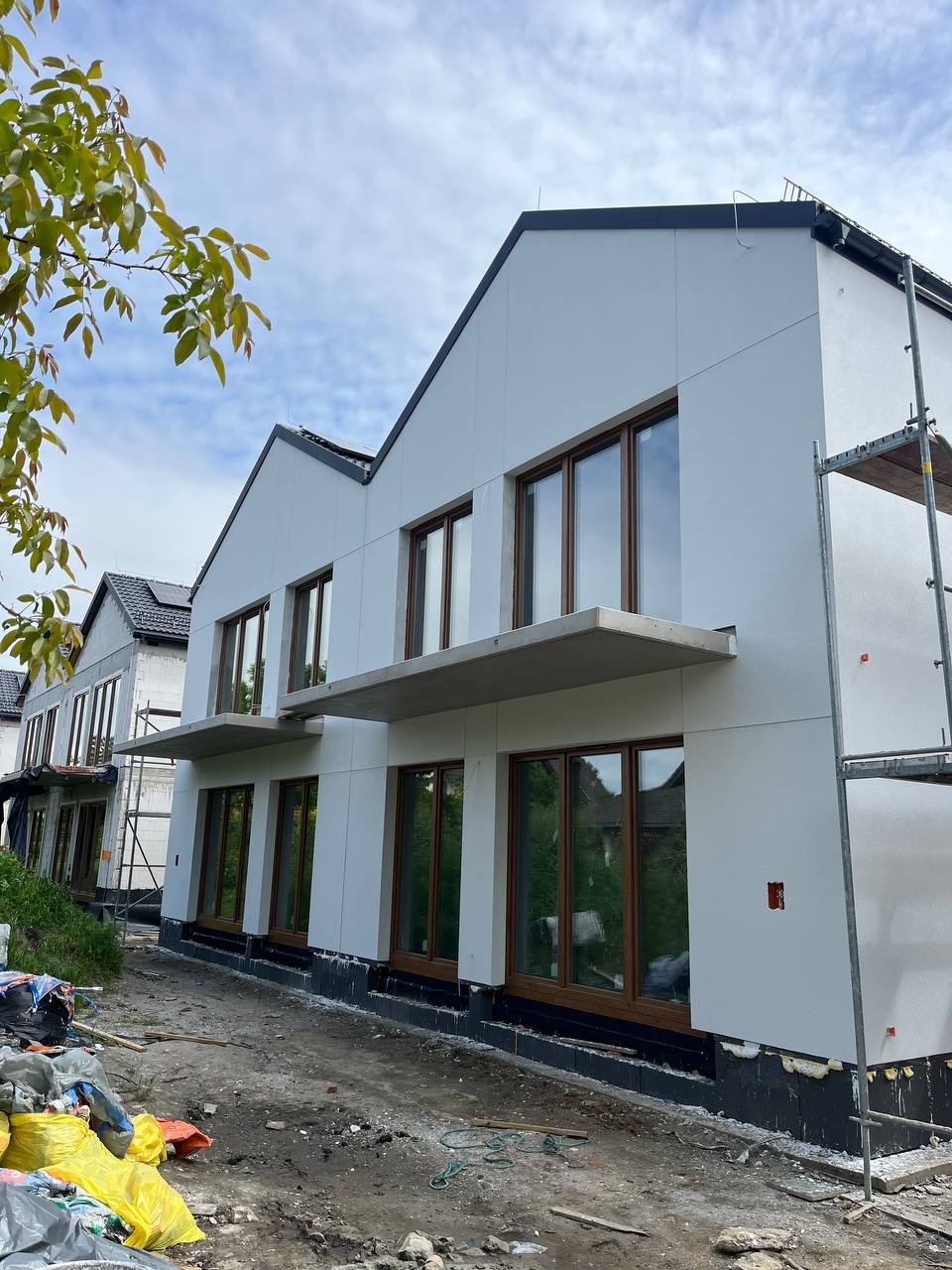 Nowoczesny dom z oknami w drewnianych ramach i świeżo otynkowaną, białą elewacją. Widoczne rusztowania i przygotowany teren wokół budynku.