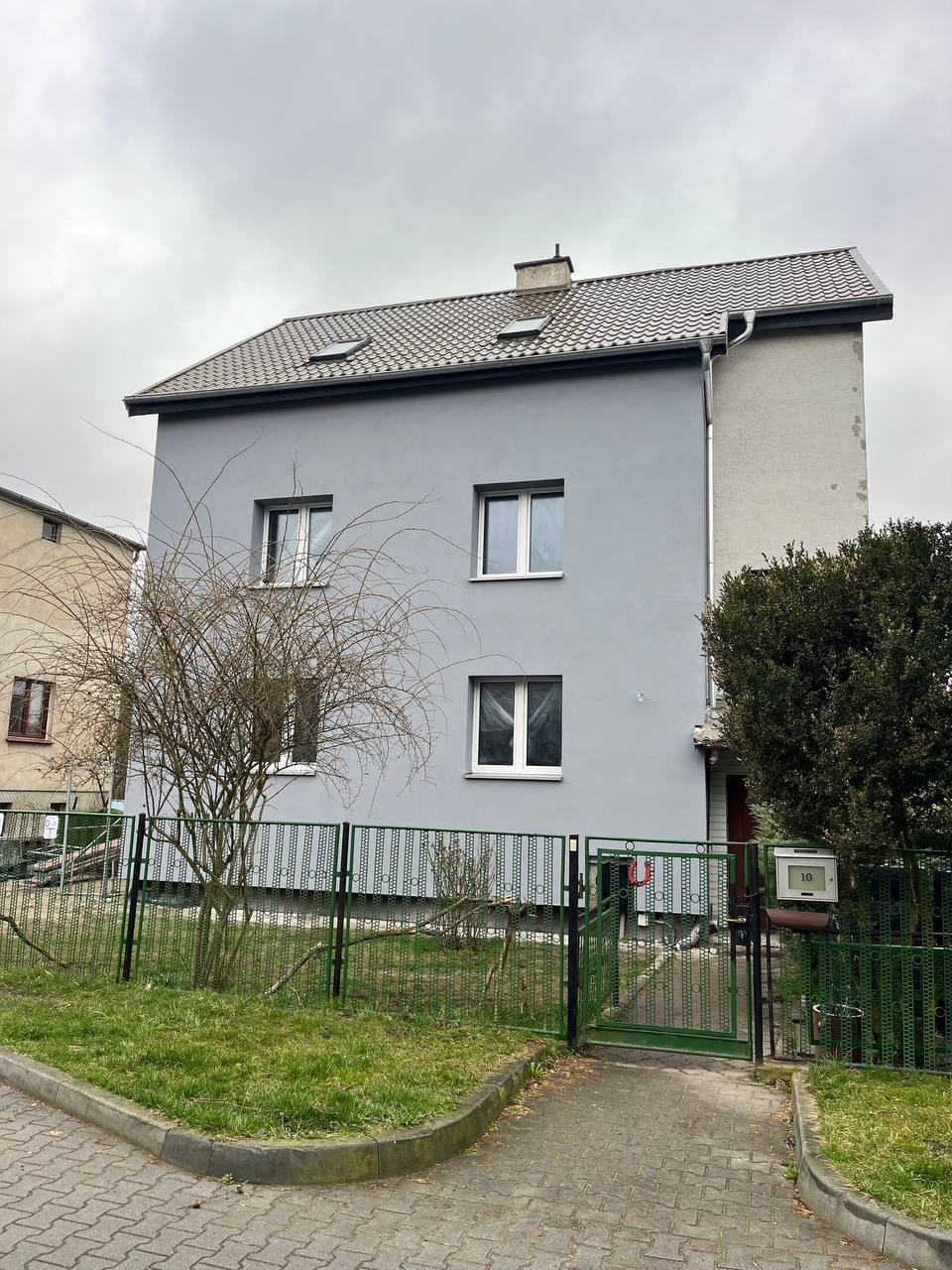 Odnowiona elewacja dwupiętrowego domu w szarym kolorze, widoczna zza zielonego ogrodzenia. Dach pokryty brązową dachówką. Pochmurne niebo w tle.