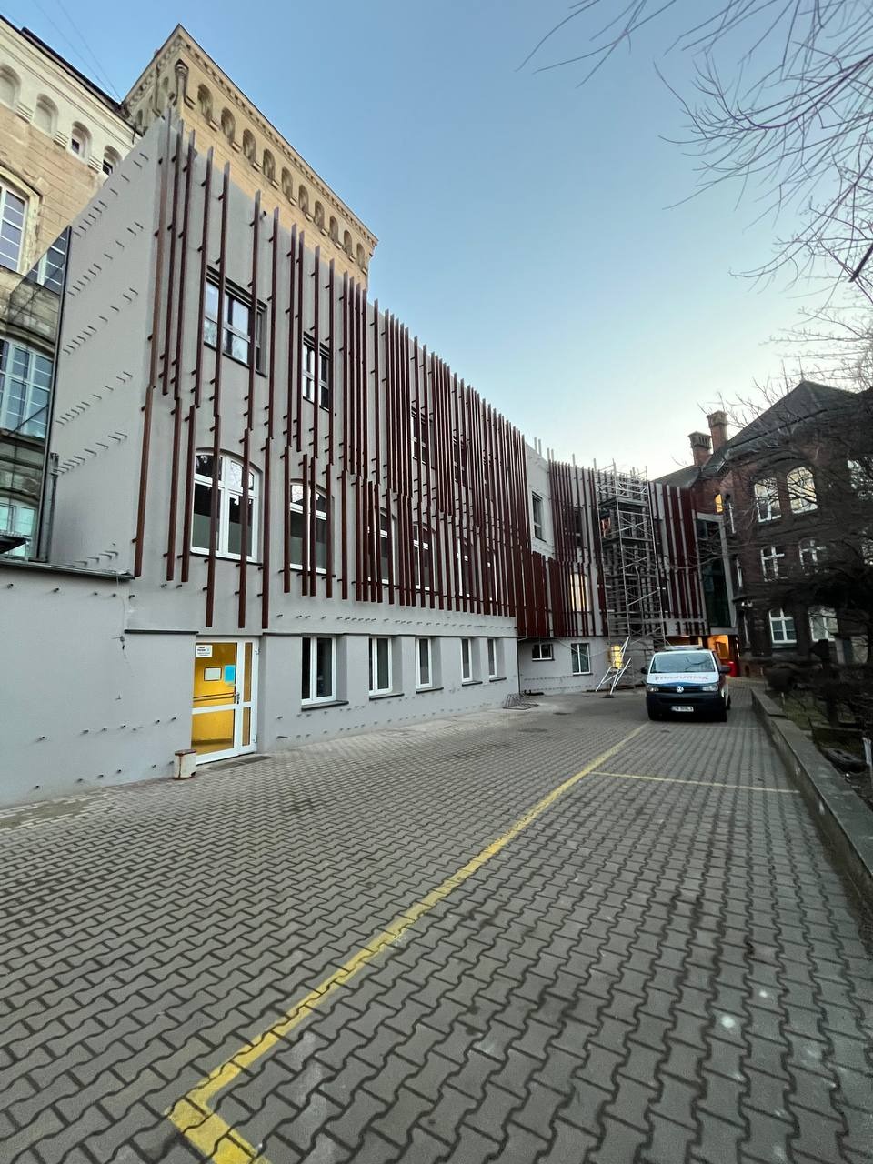 Budynek po renowacji elewacji z dekoracyjnymi, pionowymi elementami. Widoczny fragment parkingu z kostki brukowej i zaparkowany samochód dostawczy.