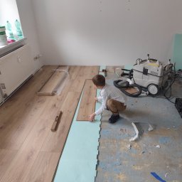 Damian Różański - Chłopiec w trakcie układania paneli podłogowych na podkładzie z pianki. Częściowo ułożona podłoga, widoczny odkurzacz przemysłowy i narzędzia.