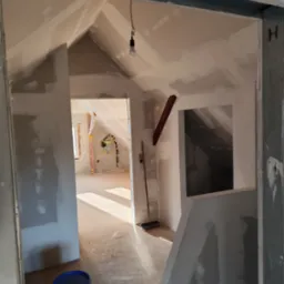 Poddasze w trakcie szpachlowania: widoczne ściany z płyt gipsowo-kartonowych, wiadro z masą szpachlową na podłodze, surowe oświetlenie, wnętrze w trakcie prac wykończeniowych.