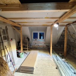 Damian Różański - Poddasze w trakcie adaptacji: widoczne drewniane belki konstrukcyjne, częściowo zamontowane płyty gipsowe, okno dachowe i materiały budowlane. Prace wykończeniowe w toku.
