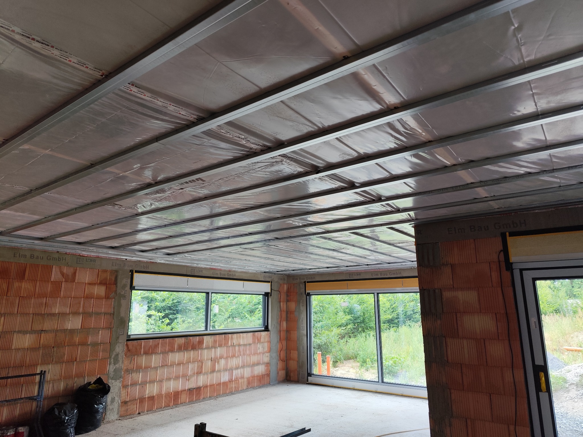 Metalowa konstrukcja pod sufitem podwieszanym w Rojewie, widoczna izolacja i ściany z cegły. W tle okna z widokiem na zieleń, pomieszczenie w stanie surowym.