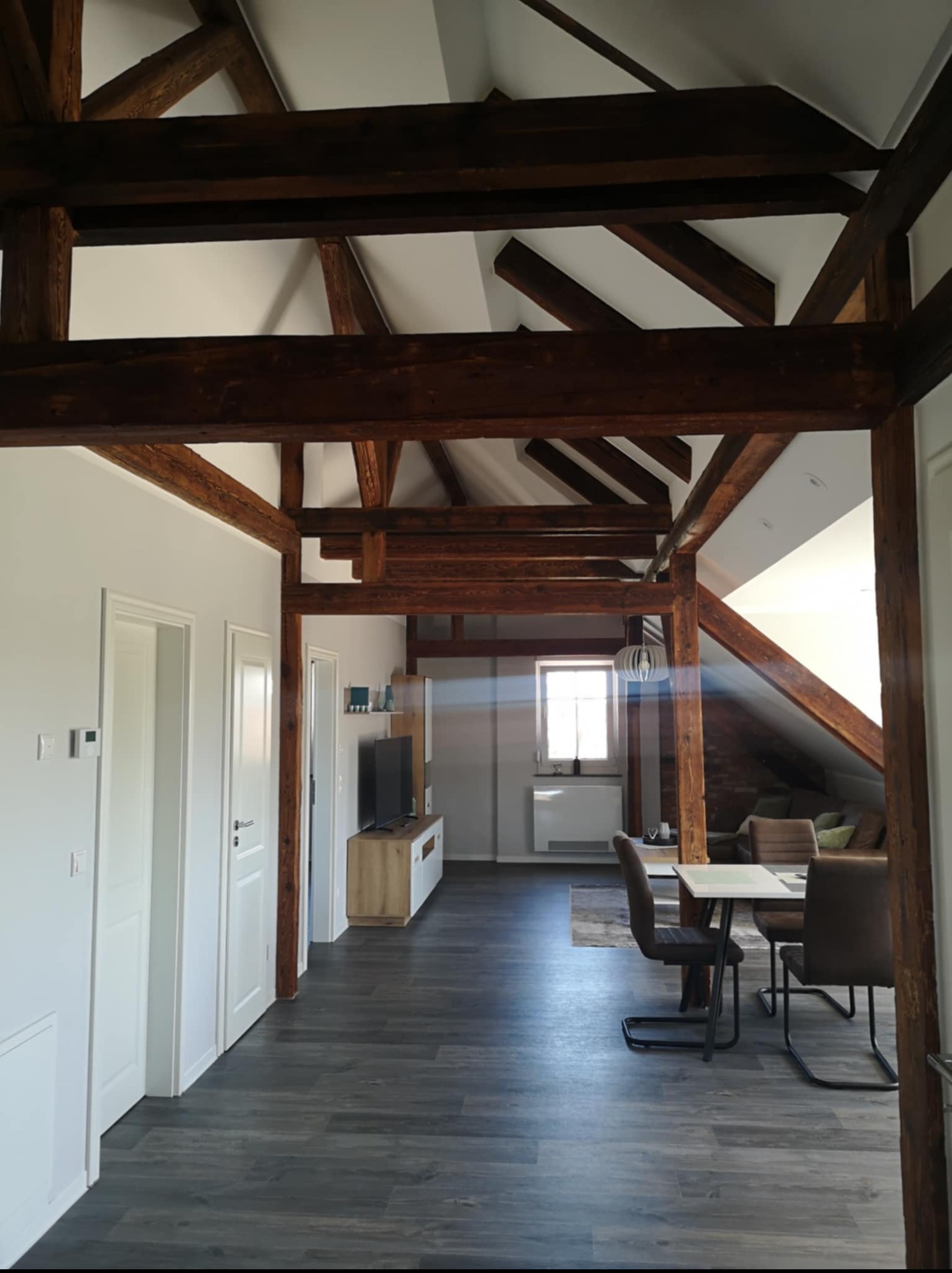Przestronne poddasze z drewnianą konstrukcją więźby dachowej, szarymi panelami podłogowymi i białymi ścianami. Widok na jadalnię i salon z oknem w tle.