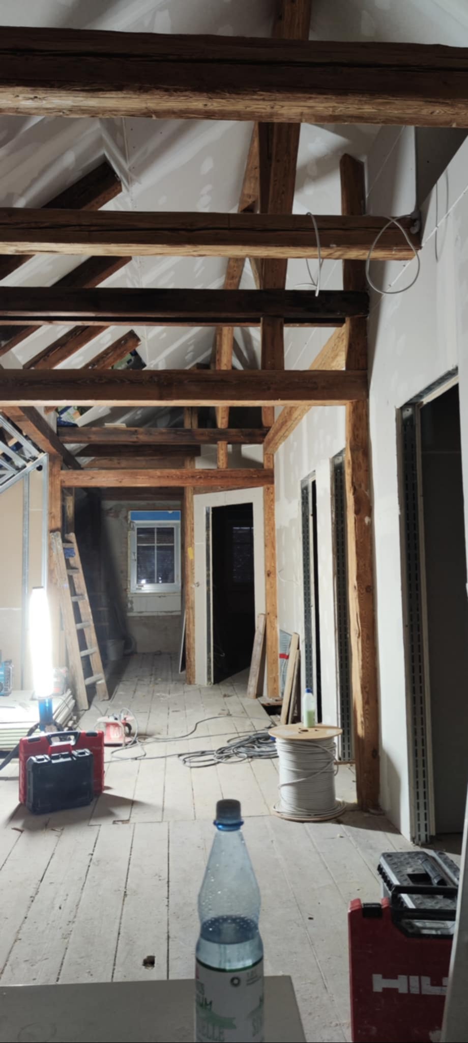 Wnętrze w trakcie remontu: surowe drewniane belki, częściowo zamontowana płyta karton-gips, narzędzia i materiały budowlane na drewnianej podłodze. Widok w głąb pomieszczeń.