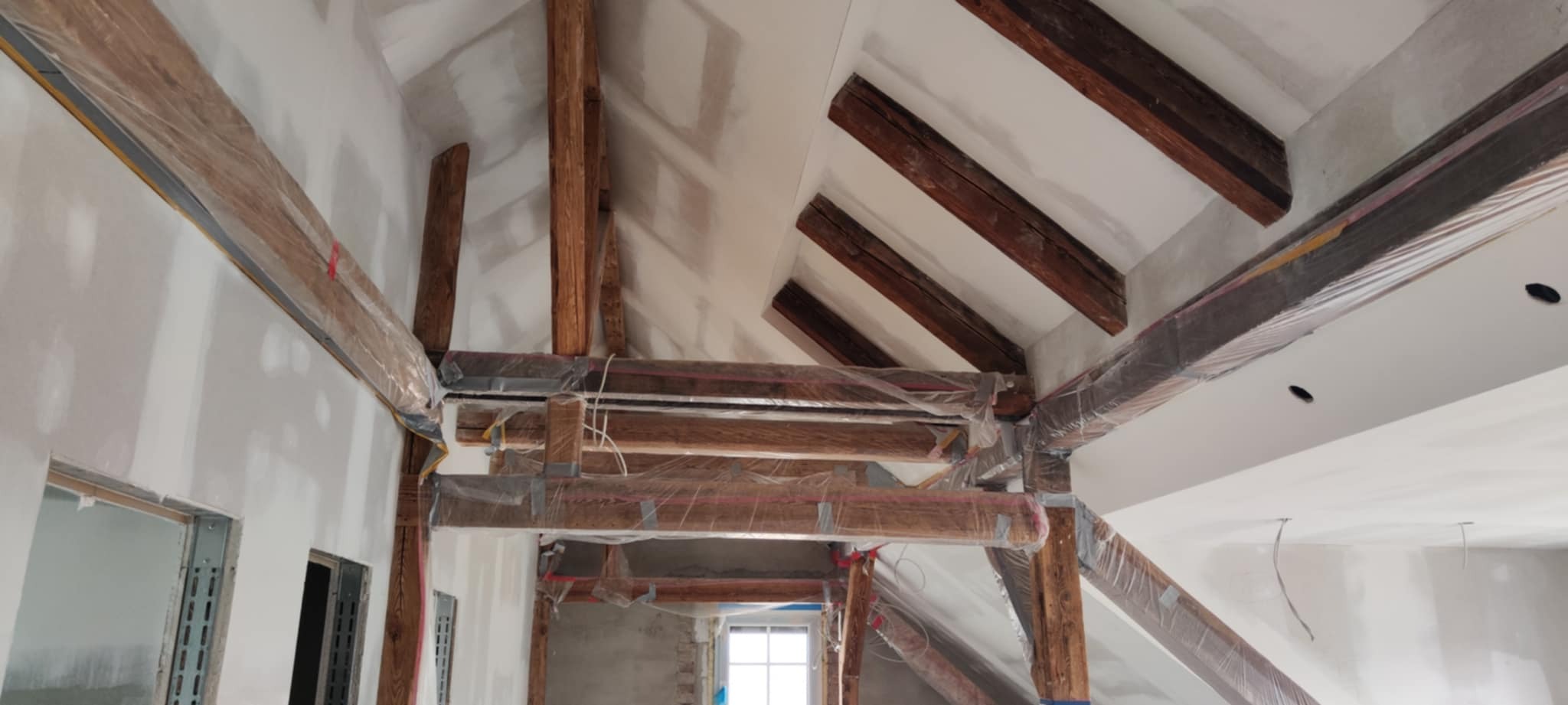 Poddasze w trakcie prac wykończeniowych: drewniane belki zabezpieczone folią, ściany pokryte gładzią gipsową, widoczne otwory okienne i sufit ze skośnymi elementami.