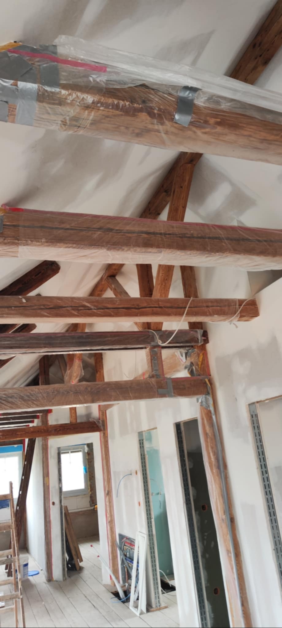Renowacja poddasza: drewniane belki stropowe zabezpieczone folią malarską, białe ściany w trakcie prac wykończeniowych, widoczne instalacje i przygotowane otwory drzwiowe.