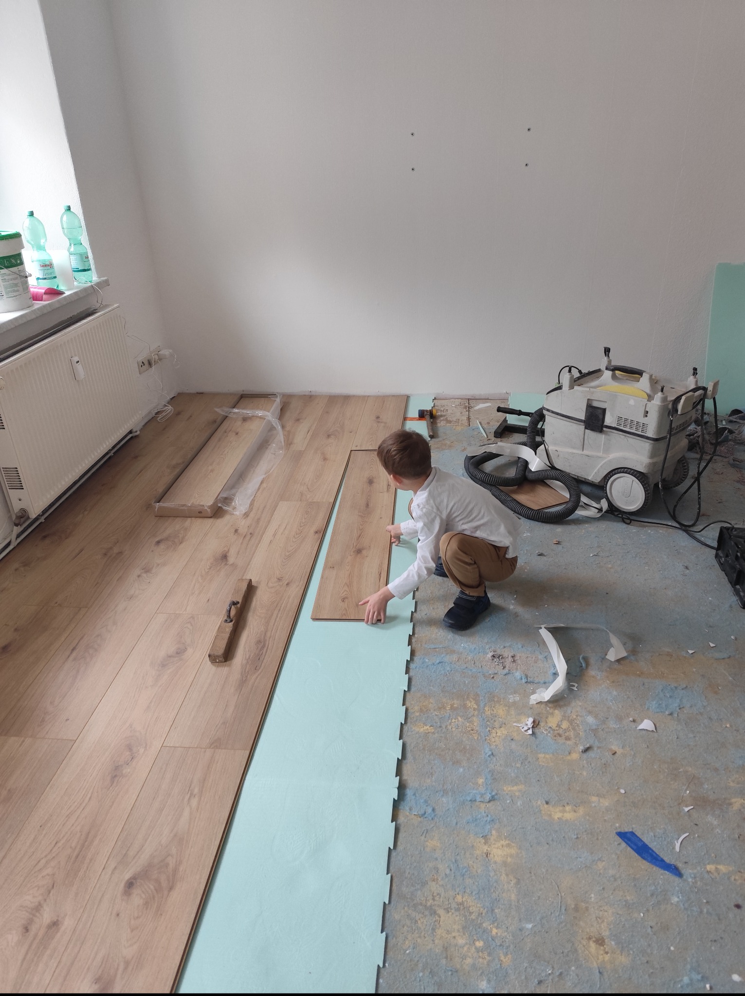 Chłopiec w trakcie układania paneli podłogowych na podkładzie z pianki. Częściowo ułożona podłoga, widoczny odkurzacz przemysłowy i narzędzia.