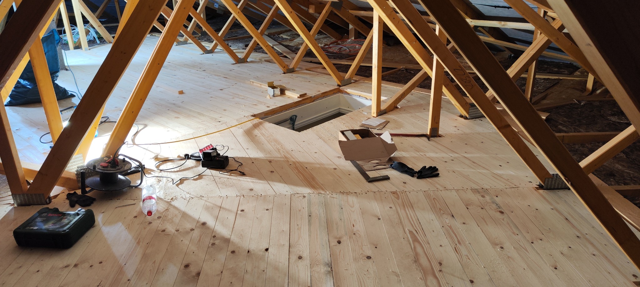 Wykończone poddasze z drewnianą podłogą, widoczne elementy konstrukcyjne dachu, narzędzia pracy i otwór rewizyjny. Jasne oświetlenie podkreśla strukturę drewna.