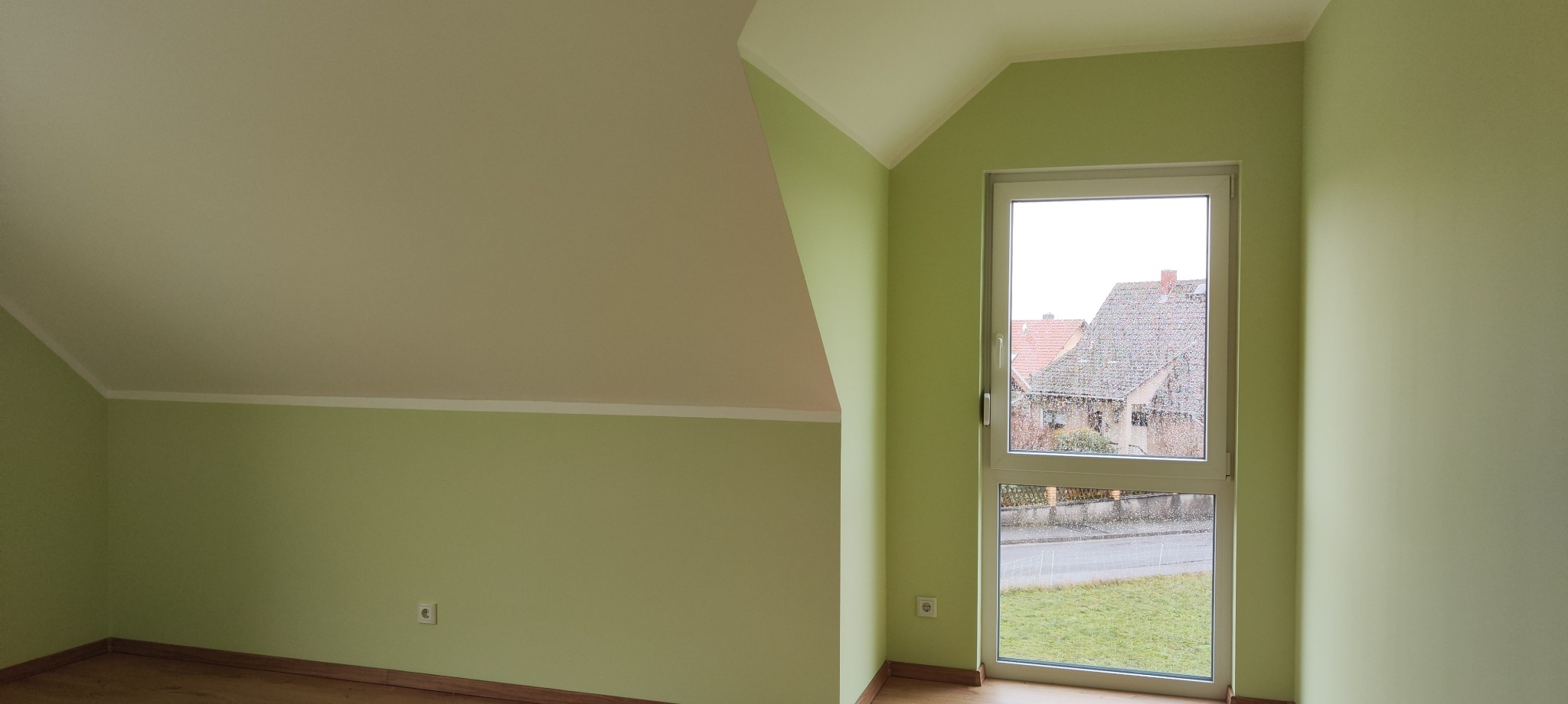 Jasne wnętrze poddasza po remoncie: zielone ściany, biały sufit, okno z widokiem na domy. Świeżo pomalowane, gładkie powierzchnie i drewniana podłoga.