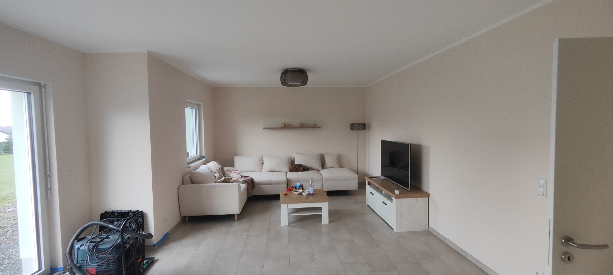 Jasne, świeżo wykończone wnętrze salonu z narożną sofą i telewizorem. Widoczny odkurzacz przemysłowy sugeruje prace wykończeniowe w toku.