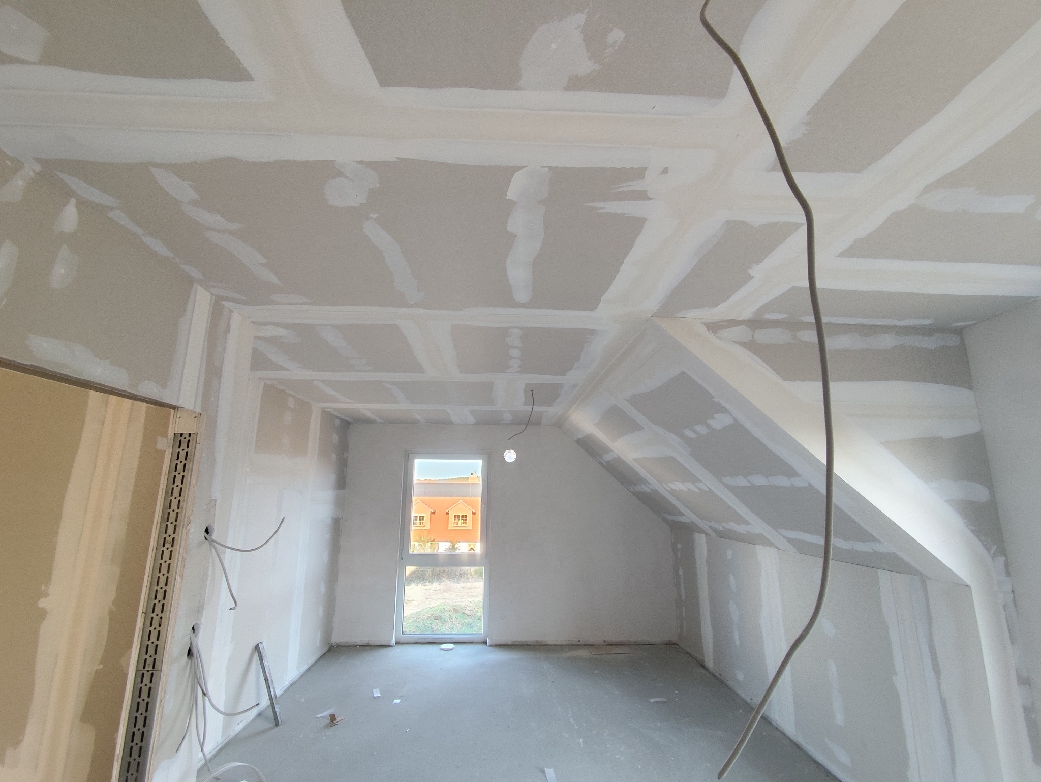 Pokój na poddaszu w trakcie prac wykończeniowych: ściany i sufit pokryte płytami kartonowo-gipsowymi, widoczne łączenia po szpachlowaniu, przewody elektryczne.