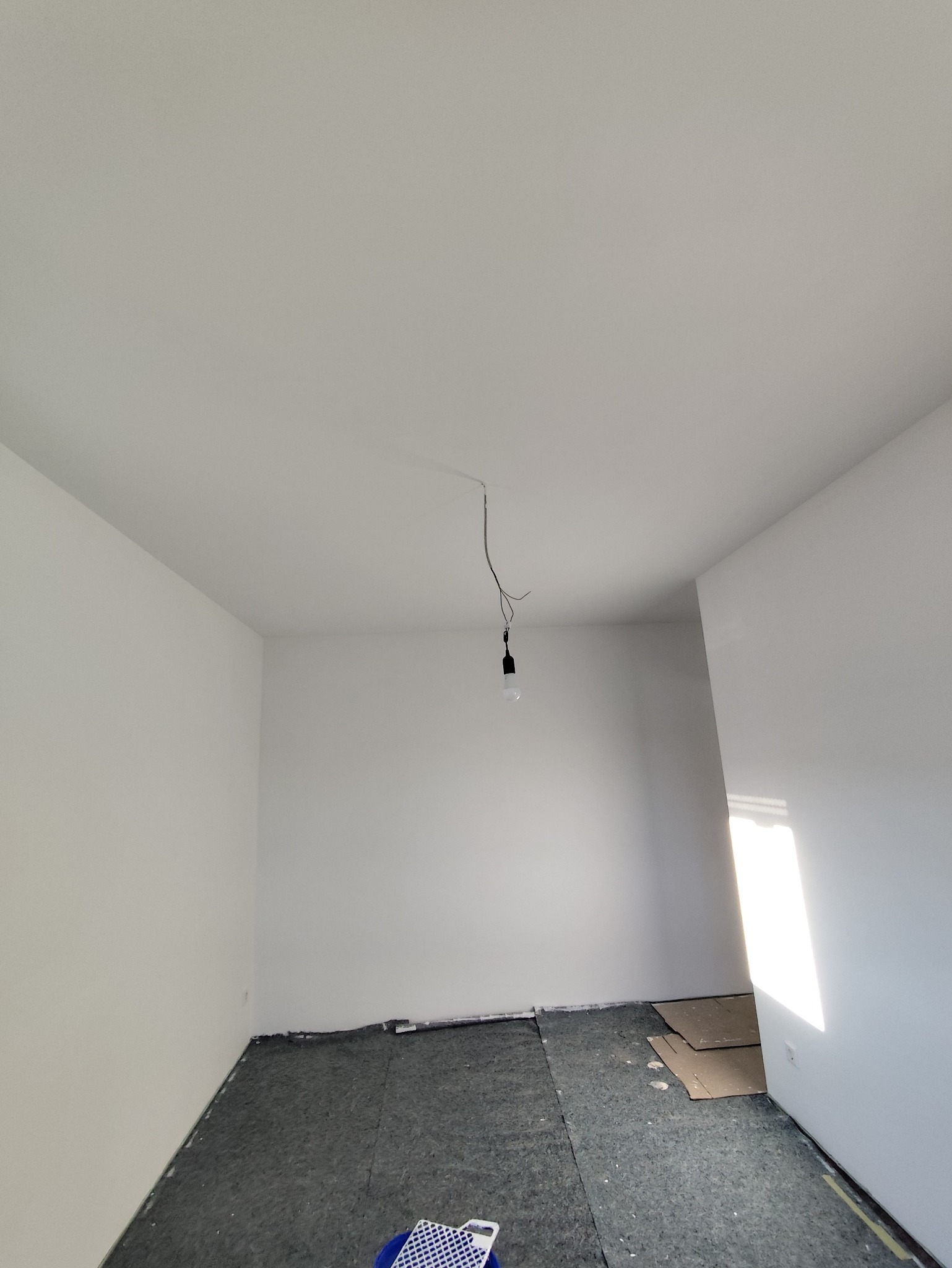Wnętrze pokoju po malowaniu na biało, widoczna podłoga zabezpieczona folią i wisząca żarówka. W rogu widoczne narzędzia malarskie.