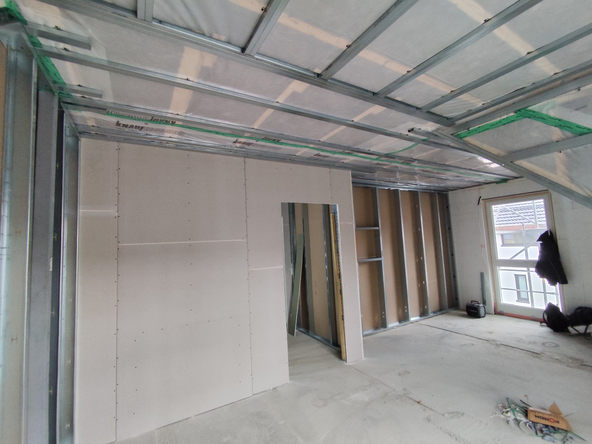 Montaż ścian działowych z płyt gipsowo-kartonowych na poddaszu. Widoczne metalowe profile i płyty Knauf. Okno w tle. Etap prac wykończeniowych.