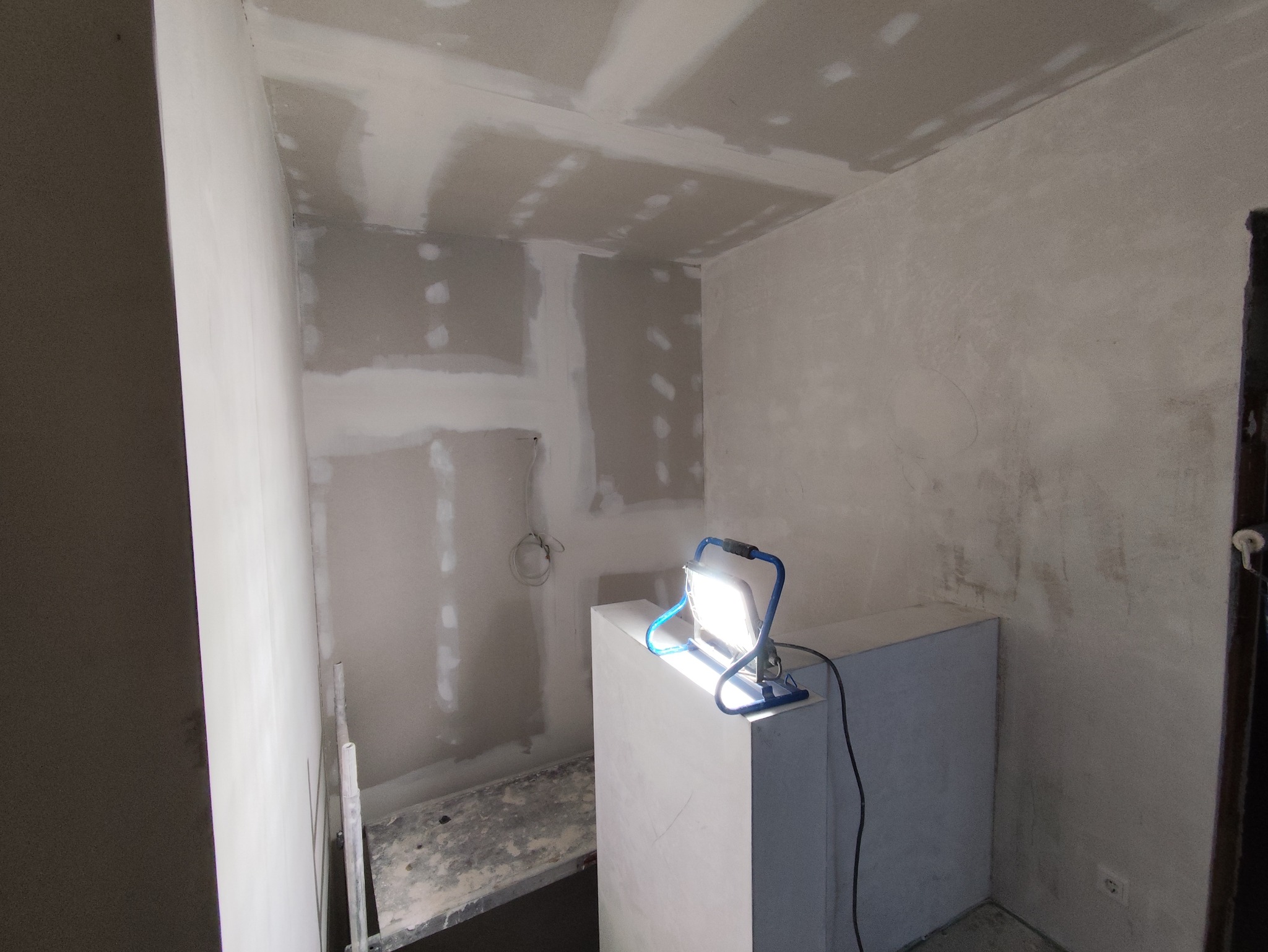 Wnętrze w trakcie prac wykończeniowych: ściany z płyt gipsowo-kartonowych, częściowo wyszpachlowane, oświetlone lampą budowlaną. Widoczny fragment drabiny.