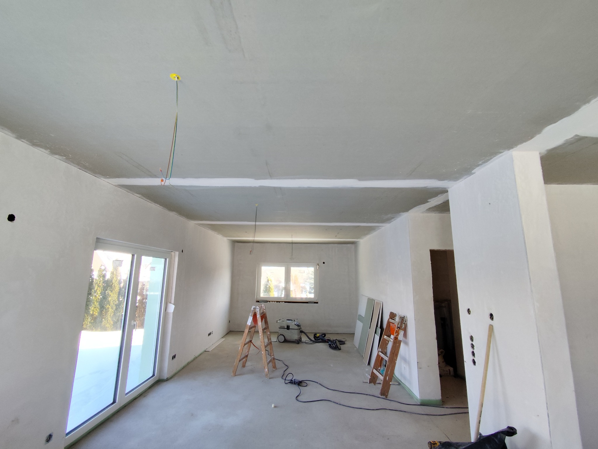 Wnętrze w trakcie szpachlowania: gładkie ściany, sufit z widocznymi przewodami, drabiny i narzędzia. Widoczne okno i drzwi balkonowe. Jasne, naturalne światło.