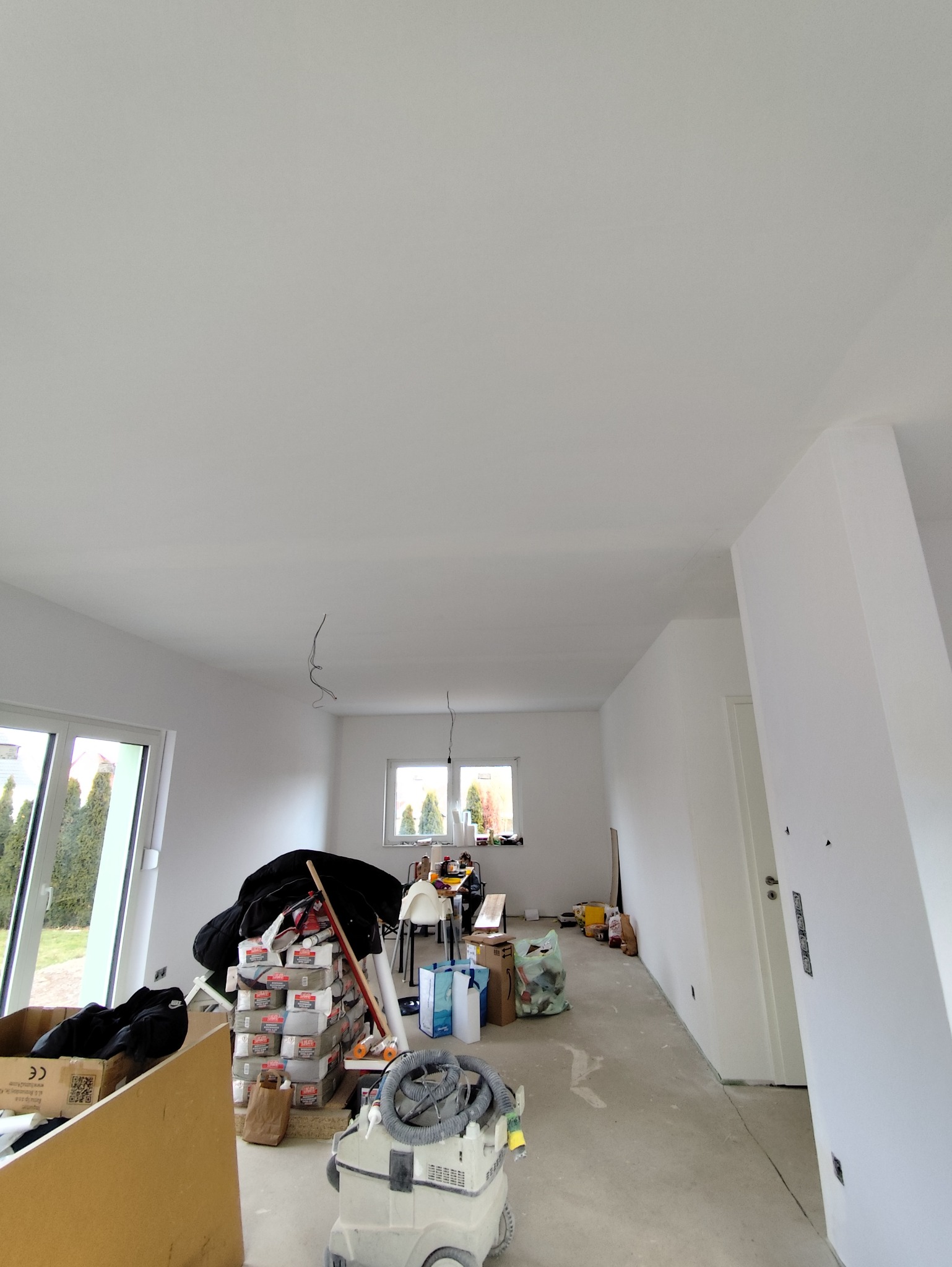 Wnętrze w trakcie prac wykończeniowych: białe ściany, surowa podłoga, materiały budowlane i odkurzacz przemysłowy. Widoczne okna i drzwi.