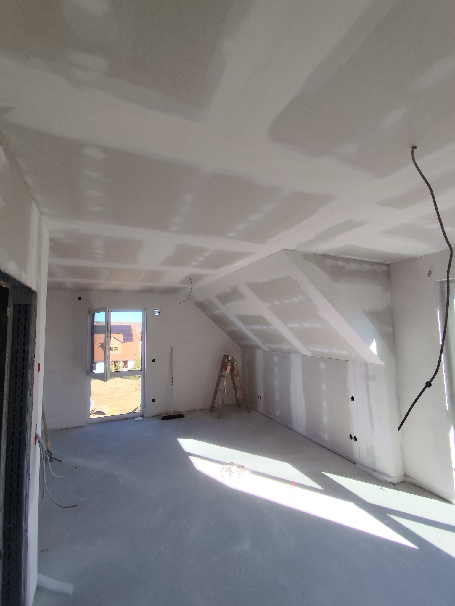 Pokój na poddaszu w trakcie prac wykończeniowych: ściany i sufit pokryte płytami karton-gips, widoczna drabina, okno z widokiem na dom. Wnętrze w stanie surowym.