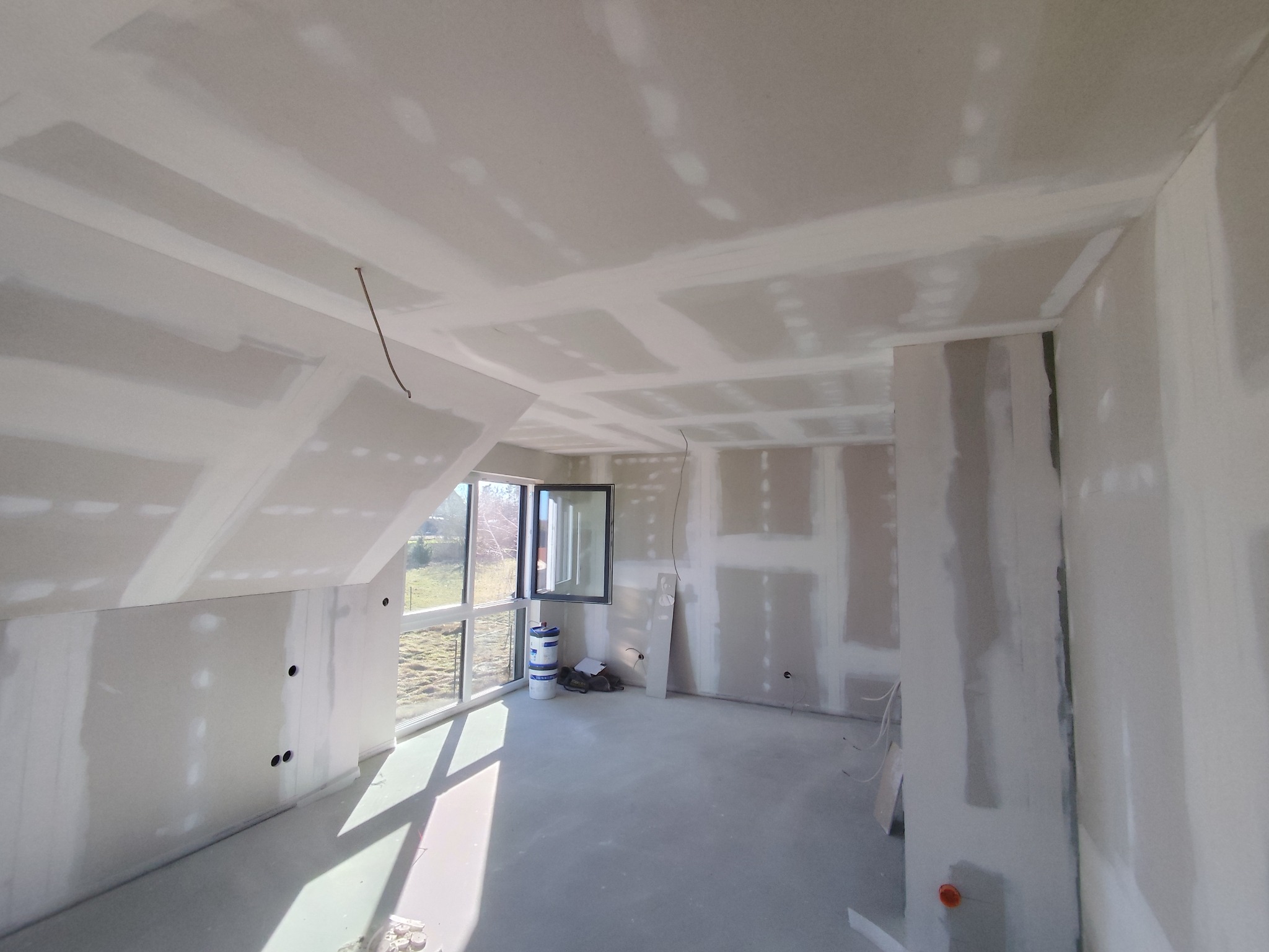 Pokój na poddaszu w trakcie prac wykończeniowych: ściany i sufit pokryte płytami kartonowo-gipsowymi, widoczne łączenia, okno z widokiem na zewnątrz, przygotowane podłoże.