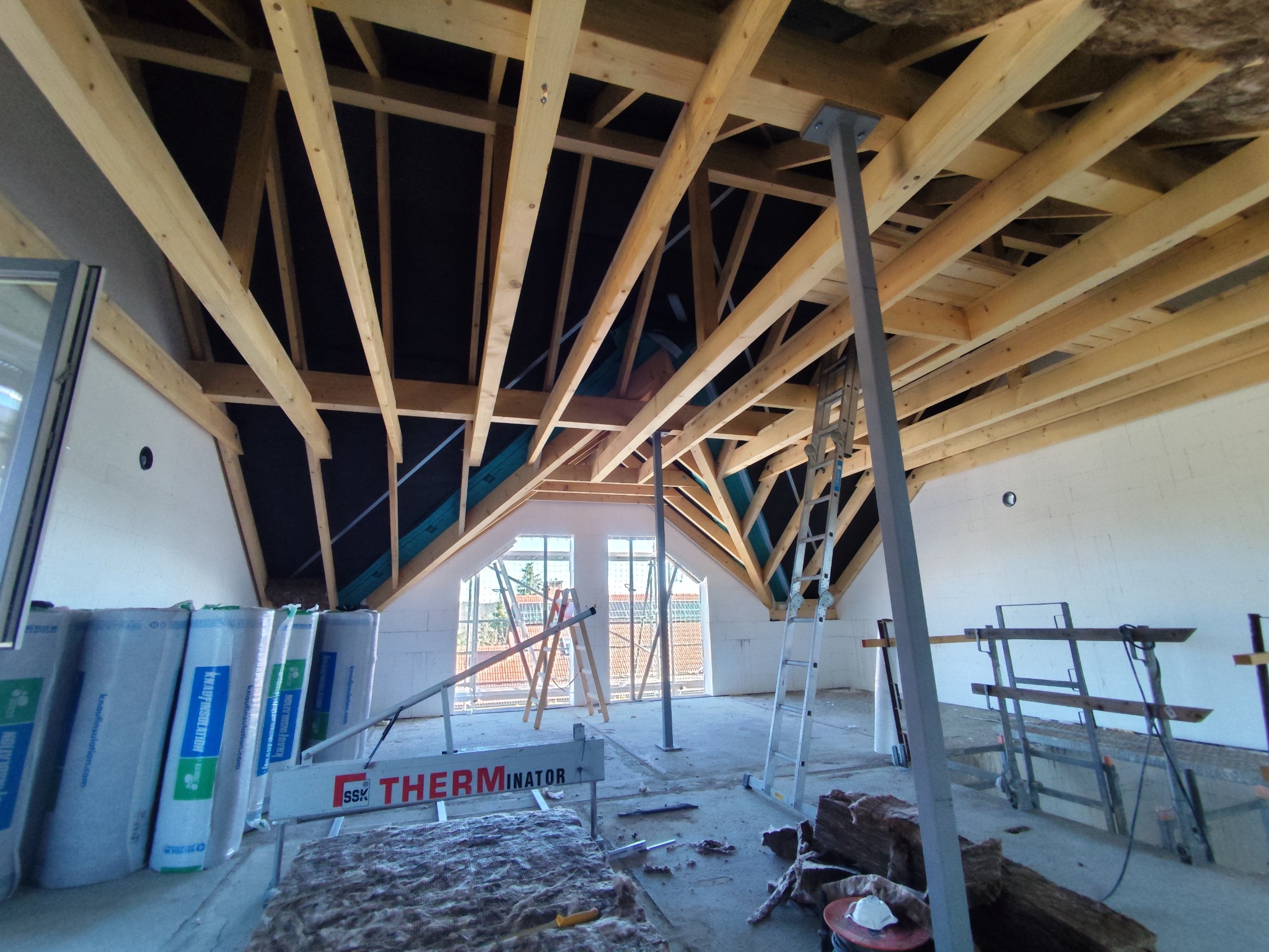 Skośny strych w trakcie prac: widoczne drewniane krokwie, izolacja, okna dachowe i rusztowania. W tle białe ściany i materiały izolacyjne.