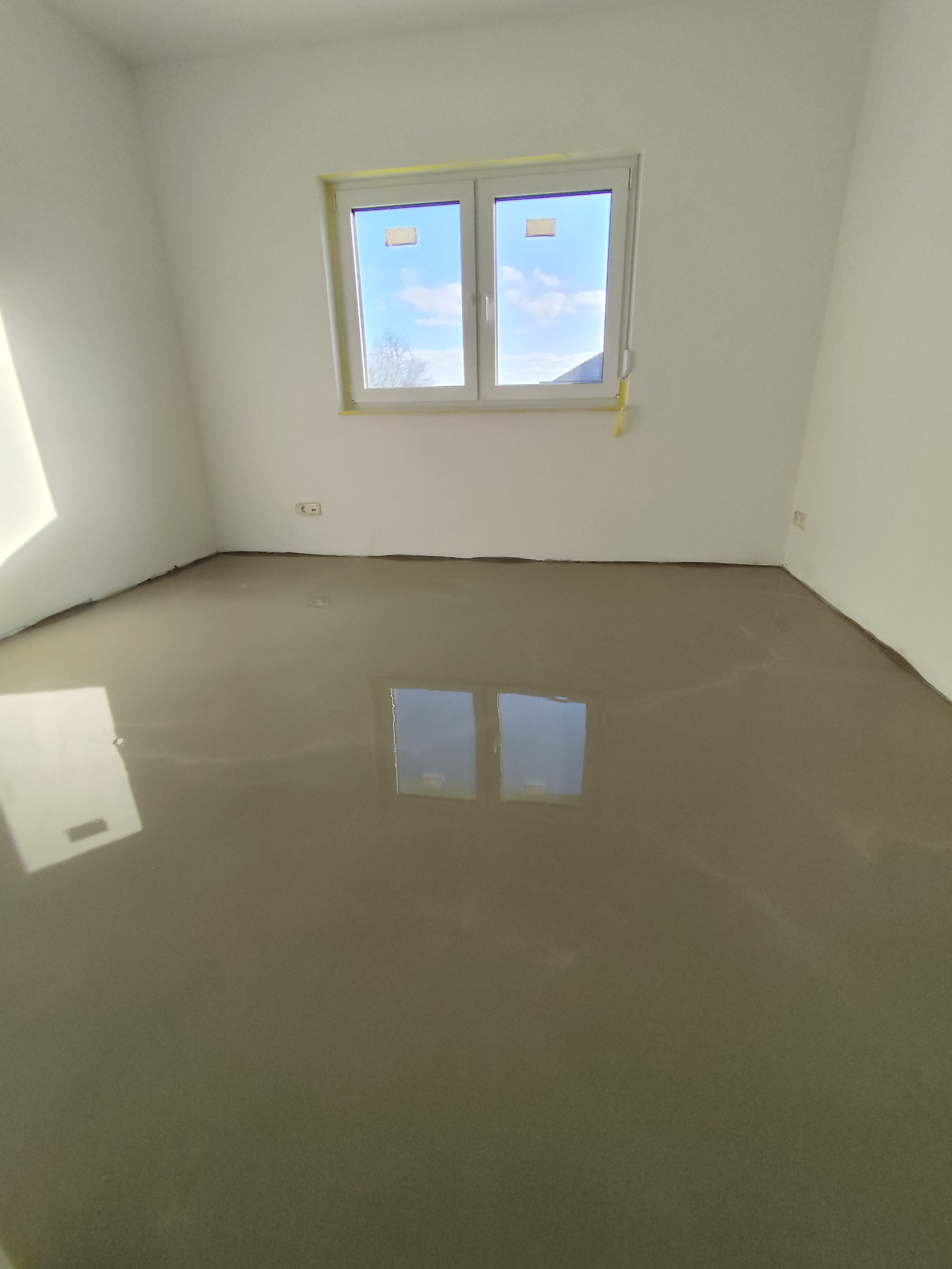 Wnętrze pokoju z wylewką samopoziomującą, okno zabezpieczone taśmą malarską. Widoczne gniazdko elektryczne. Podłoga odbija światło, tworząc lustrzany efekt.