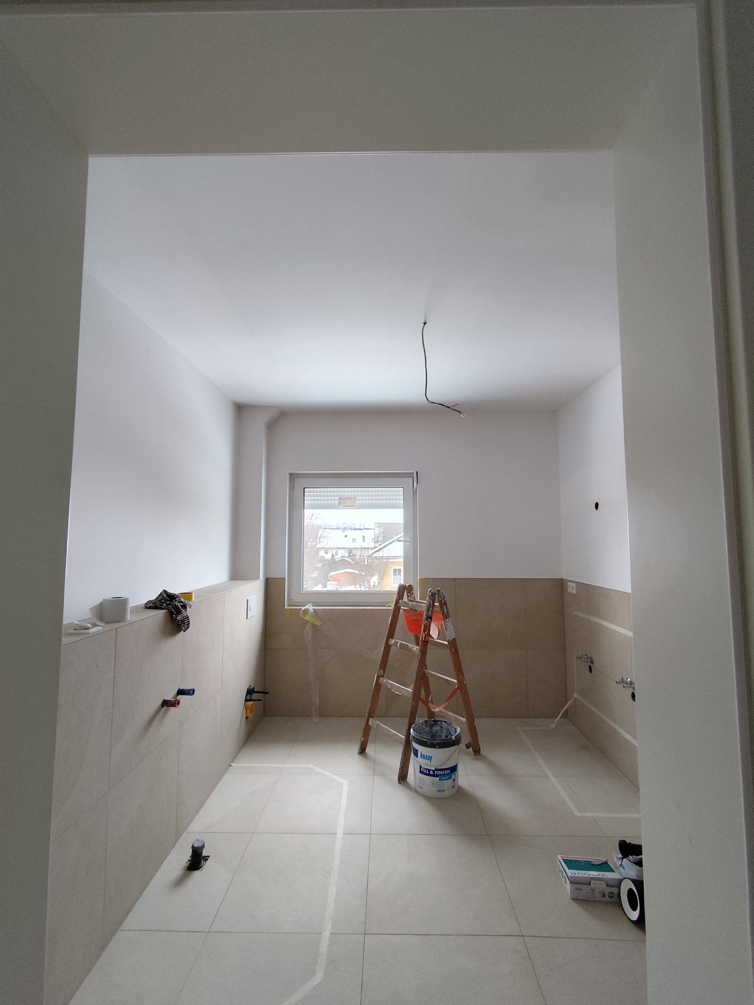 Wnętrze łazienki w trakcie prac wykończeniowych: widoczna drabina, wiadro z farbą, zaznaczone obszary na podłodze, płytki na ścianach i wystające rury.