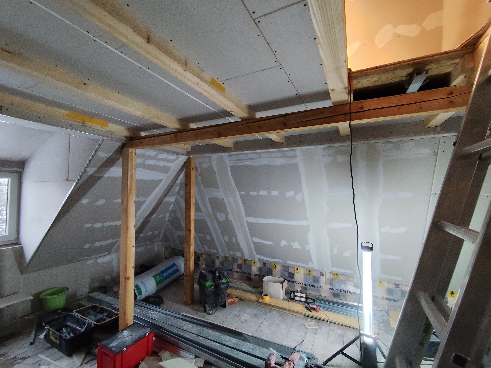 Skośne poddasze w trakcie adaptacji: drewniana konstrukcja, płyty karton-gips na ścianach, narzędzia i materiały budowlane. Widoczne elementy instalacji elektrycznej.