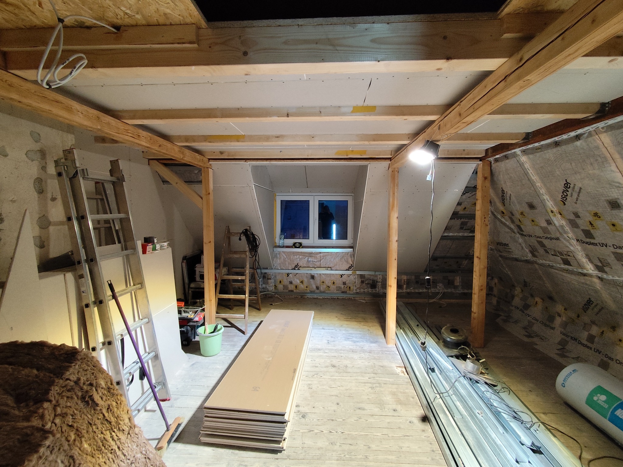 Poddasze w trakcie adaptacji: widoczne drewniane belki konstrukcyjne, częściowo zamontowane płyty gipsowe, okno dachowe i materiały budowlane. Prace wykończeniowe w toku.