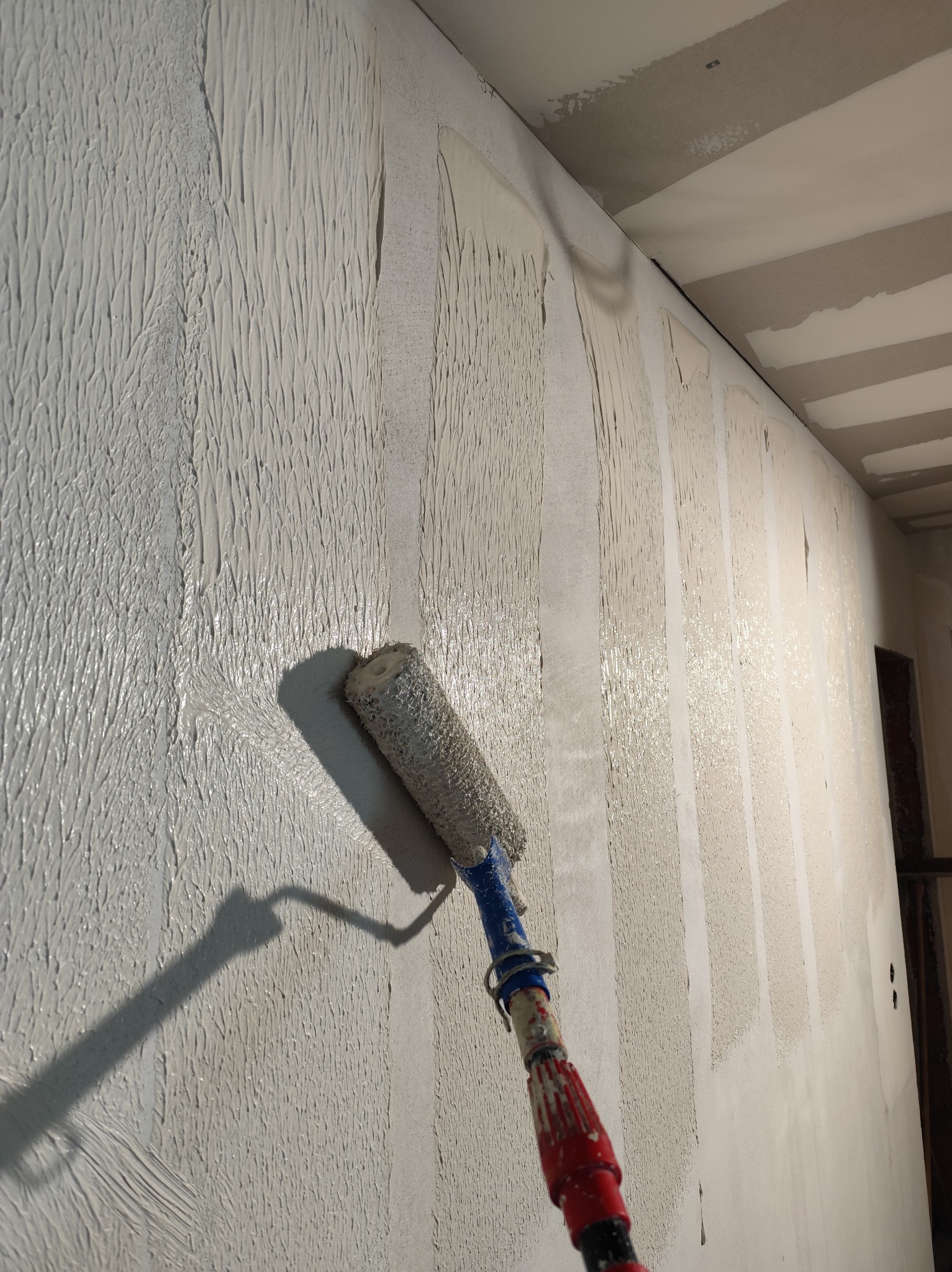 Świeżo nałożona, gruba warstwa szpachli na ścianie z widocznymi śladami wałka malarskiego. W tle sufit z płyt kartonowo-gipsowych i fragment ościeżnicy drzwi.