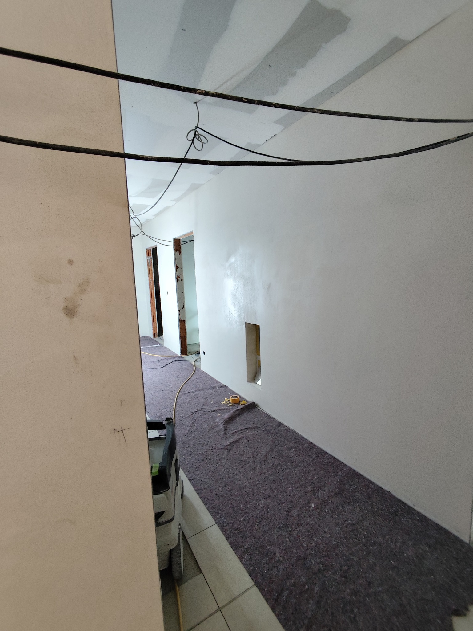 Białe ściany po szpachlowaniu w korytarzu, zabezpieczona podłoga, widoczne kable i otwory w ścianie. Prace wykończeniowe w trakcie remontu mieszkania.