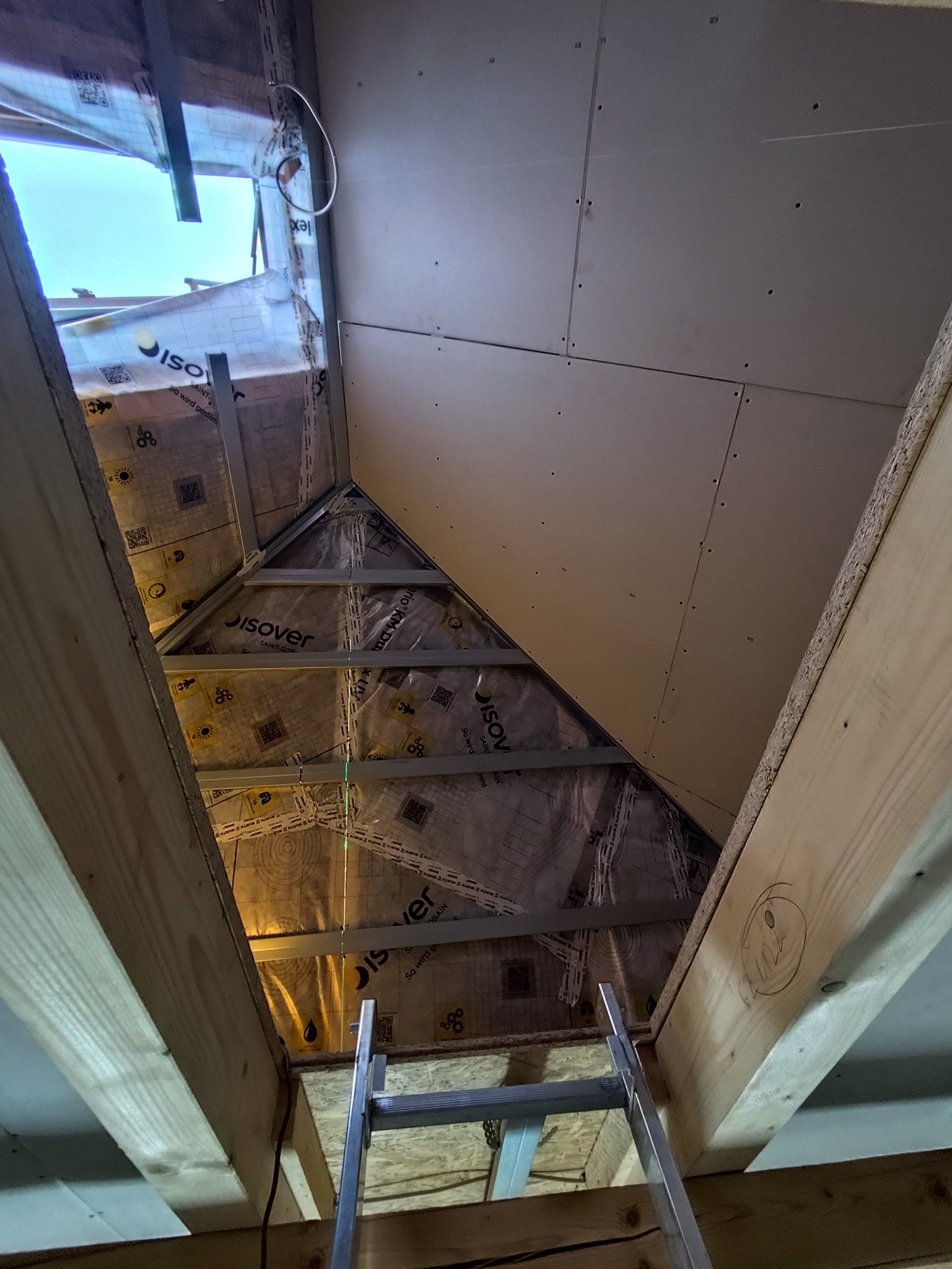 Skośny strych z izolacją i częściowo zamontowaną płytą gipsową, widok z dołu na drabinę. Widoczna folia izolacyjna z nadrukiem i metalowe profile.