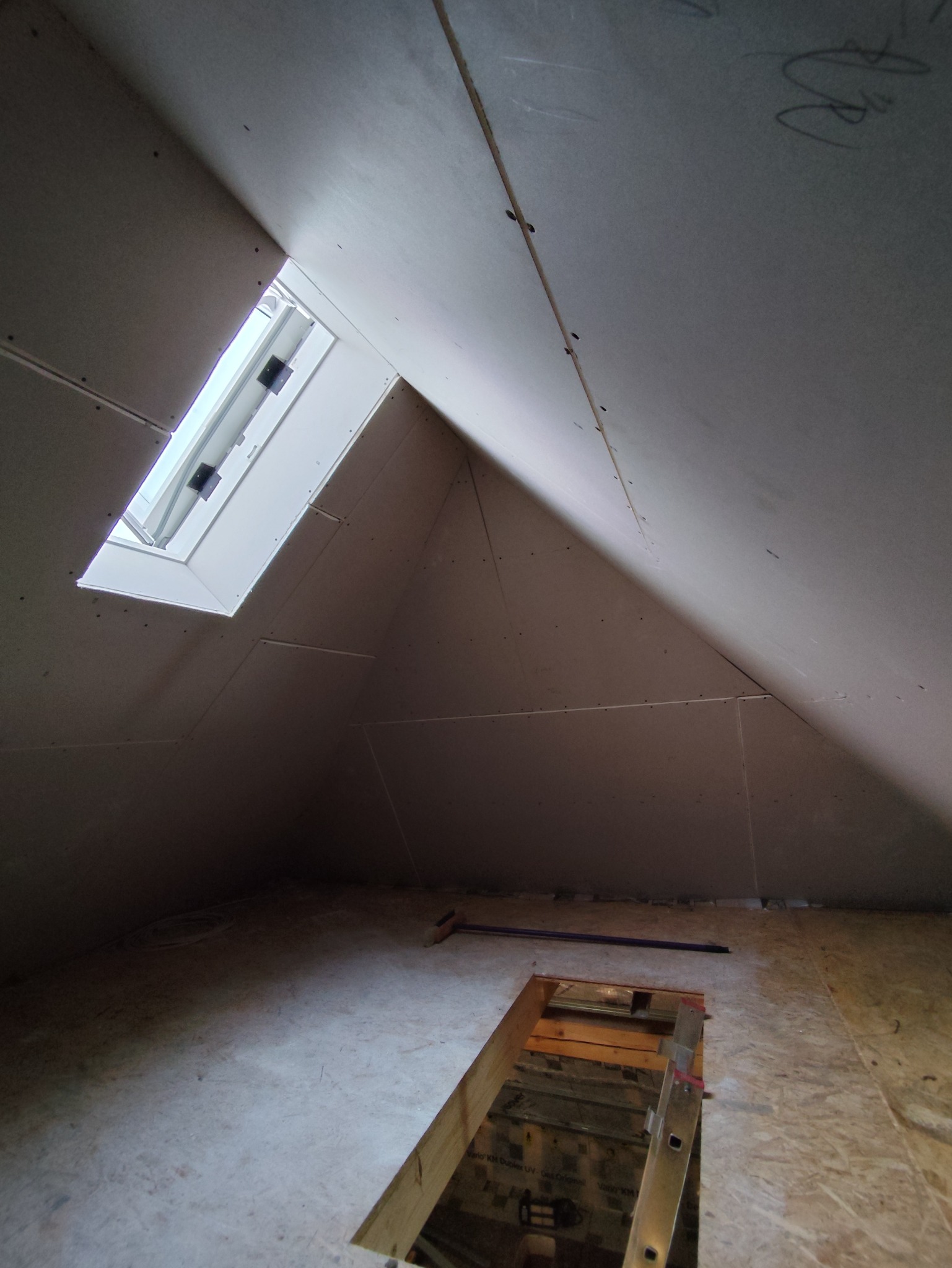 Poddasze z oknem dachowym w trakcie montażu płyt karton-gips, widoczne łączenia i mocowania. Na podłodze właz i drabina.