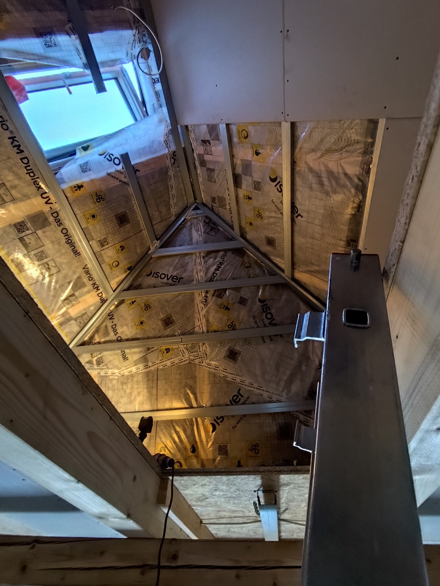 Skośny sufit poddasza w trakcie izolacji, widoczne metalowe profile, folia izolacyjna z nadrukiem i fragment okna dachowego. Prace wykończeniowe na poddaszu.