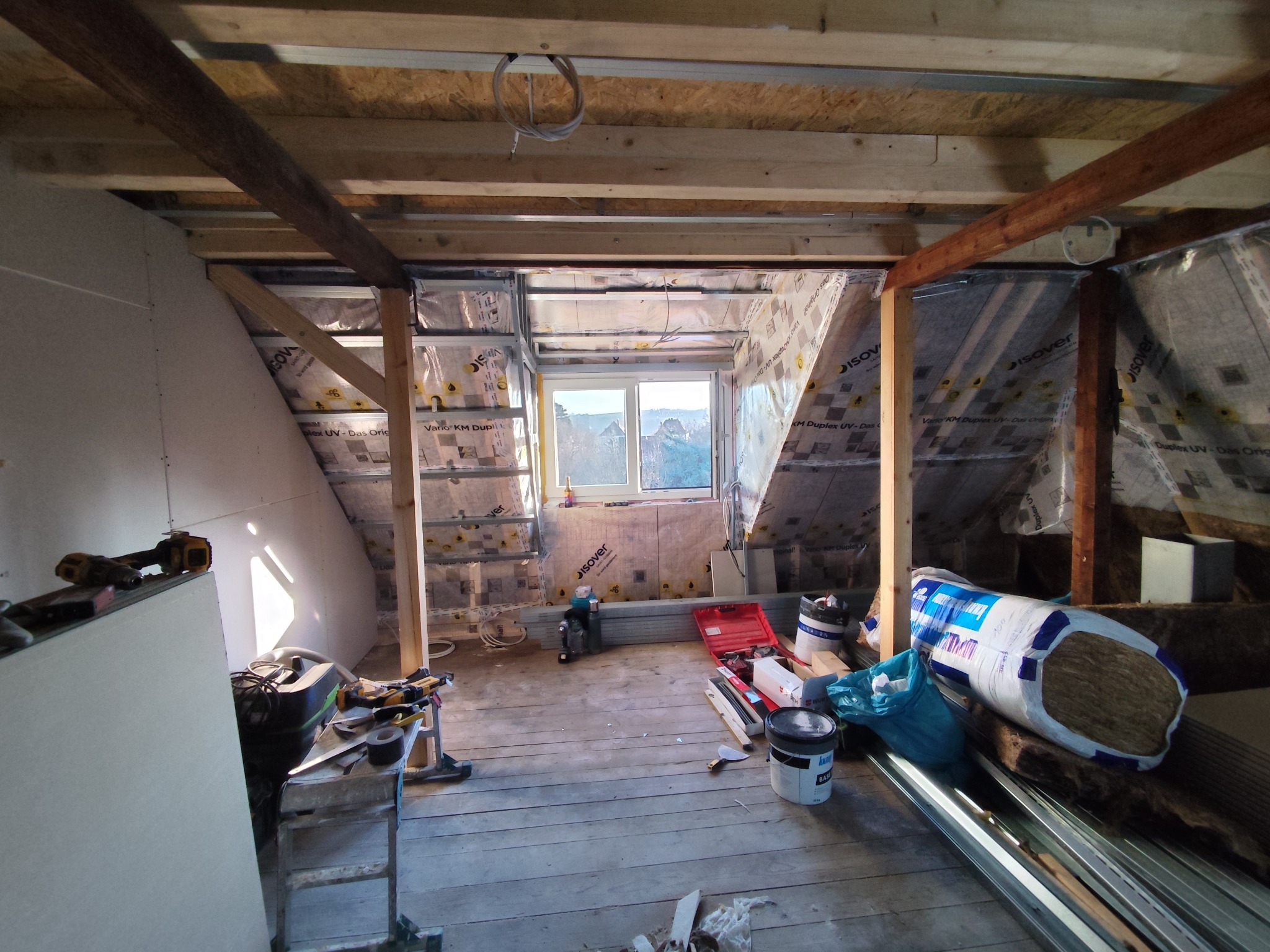 Adaptacja poddasza: widok na ocieplone ściany skośne z oknem, konstrukcja z profili, narzędzia i materiały budowlane na drewnianej podłodze. Etap prac wykończeniowych.