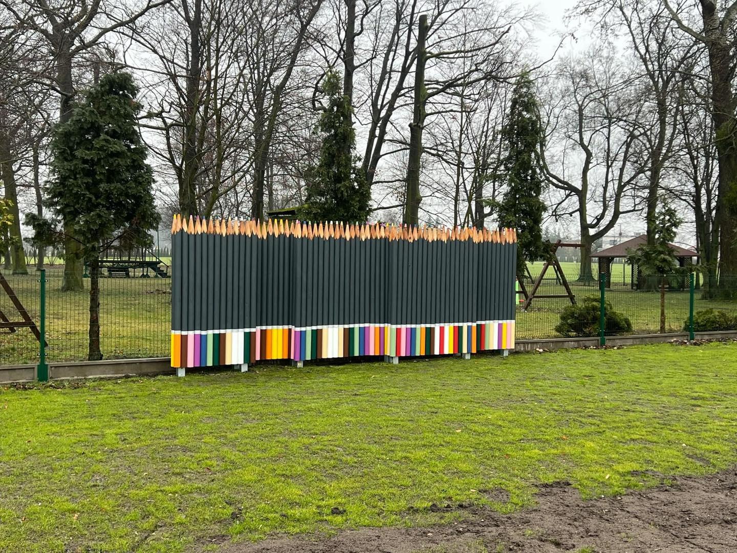 Nietypowe ogrodzenie w formie kolorowych kredek na tle parku. Ciemne kredki u góry, barwne paski na dole. Widoczne drzewa bez liści i trawnik.