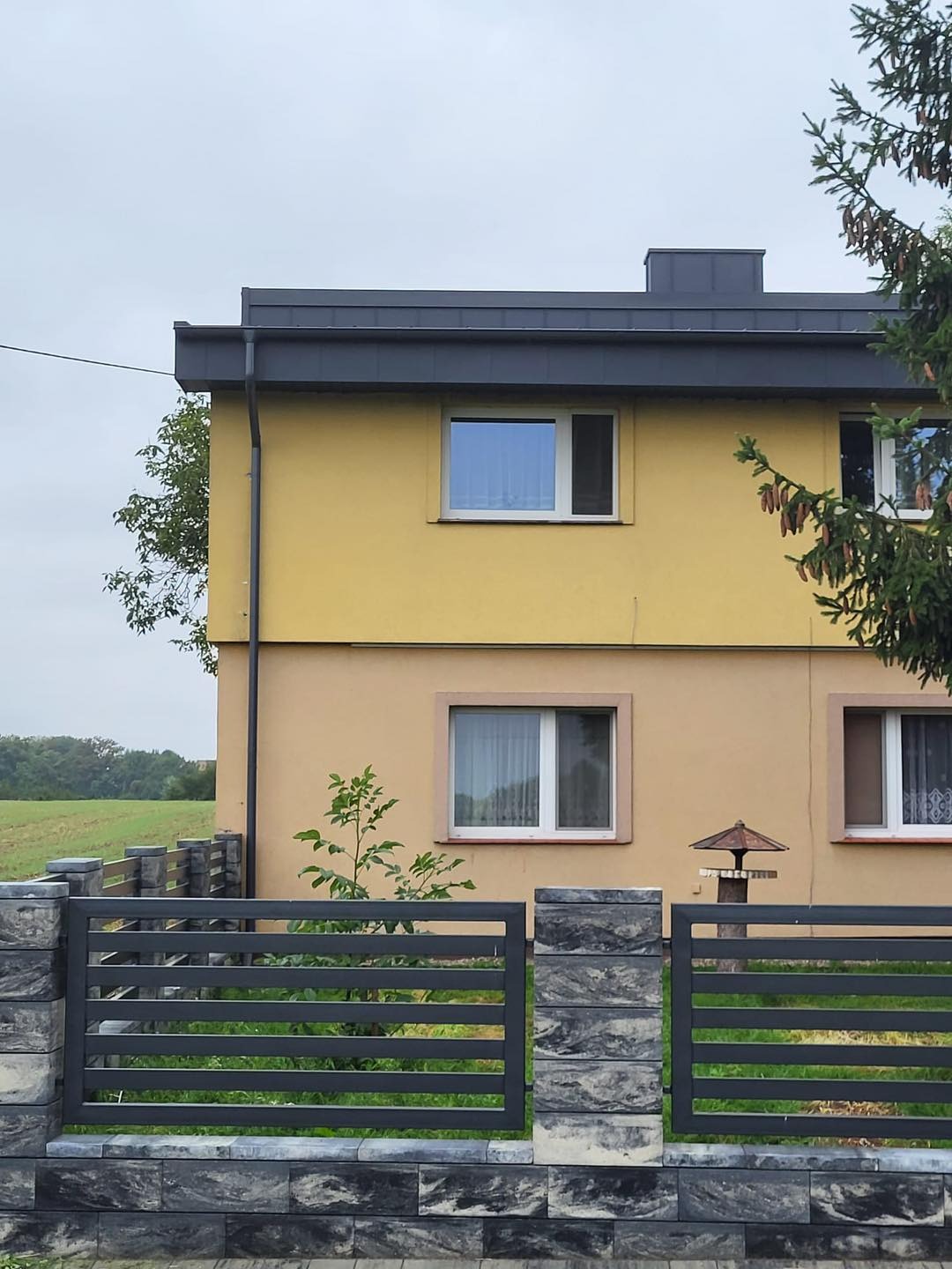 Dwukondygnacyjny dom z ciemnoszarym dachem i rynnami, widziany zza nowoczesnego ogrodzenia z szarej kostki. Tło stanowi zielone pole i pochmurne niebo.