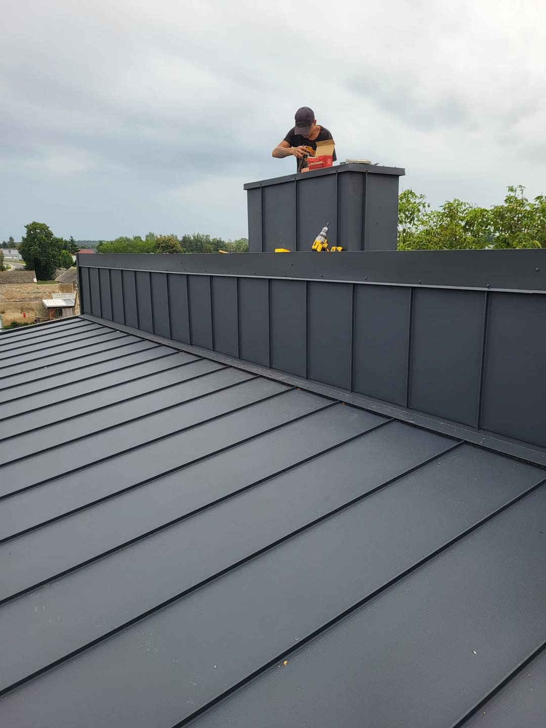 Dekarz montuje obróbkę blacharską komina na dachu z blachy na rąbek stojący. Na dachu leży wkrętarka. Widok z góry na nowy, szary dach i pracę dekarza.