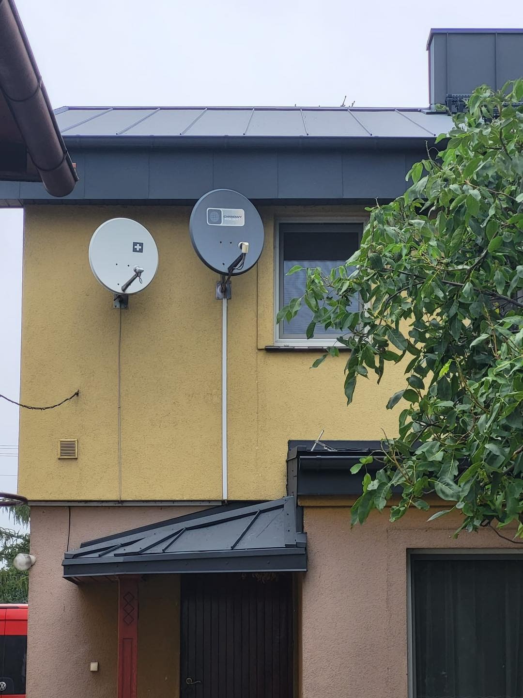 Dwukondygnacyjny budynek z szarym dachem i dwoma antenami satelitarnymi na żółtej elewacji, obok zielone drzewo i szara blacha nad wejściem.