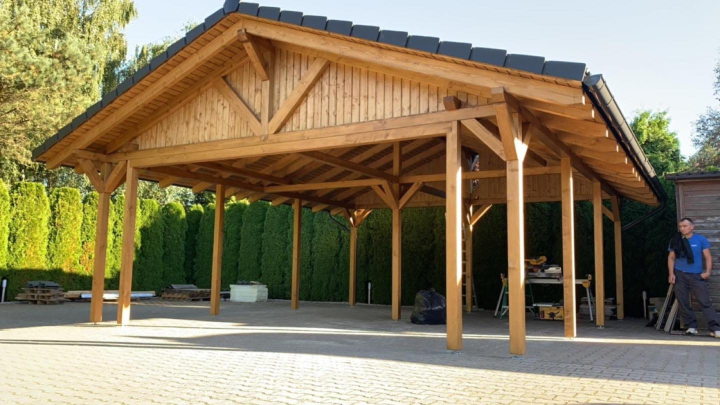 Drewniana wiata z czarną dachówką, wsparta na solidnych słupach, na tle zielonego żywopłotu. Widoczny pracownik w tle. Konstrukcja z drewna, dach kryty dachówką.