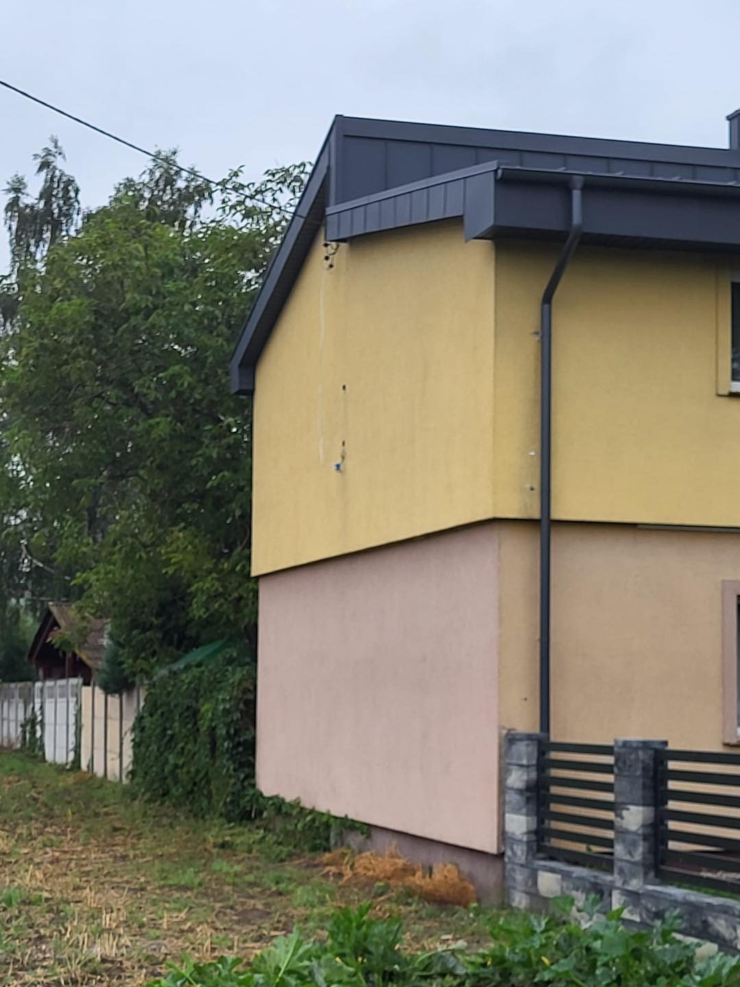 Żółto-różowy dom z ciemnoszarą blachą na dachu i rynną. Widoczny fragment ogrodzenia i zieleń.  Ujęcie z poziomu gruntu.