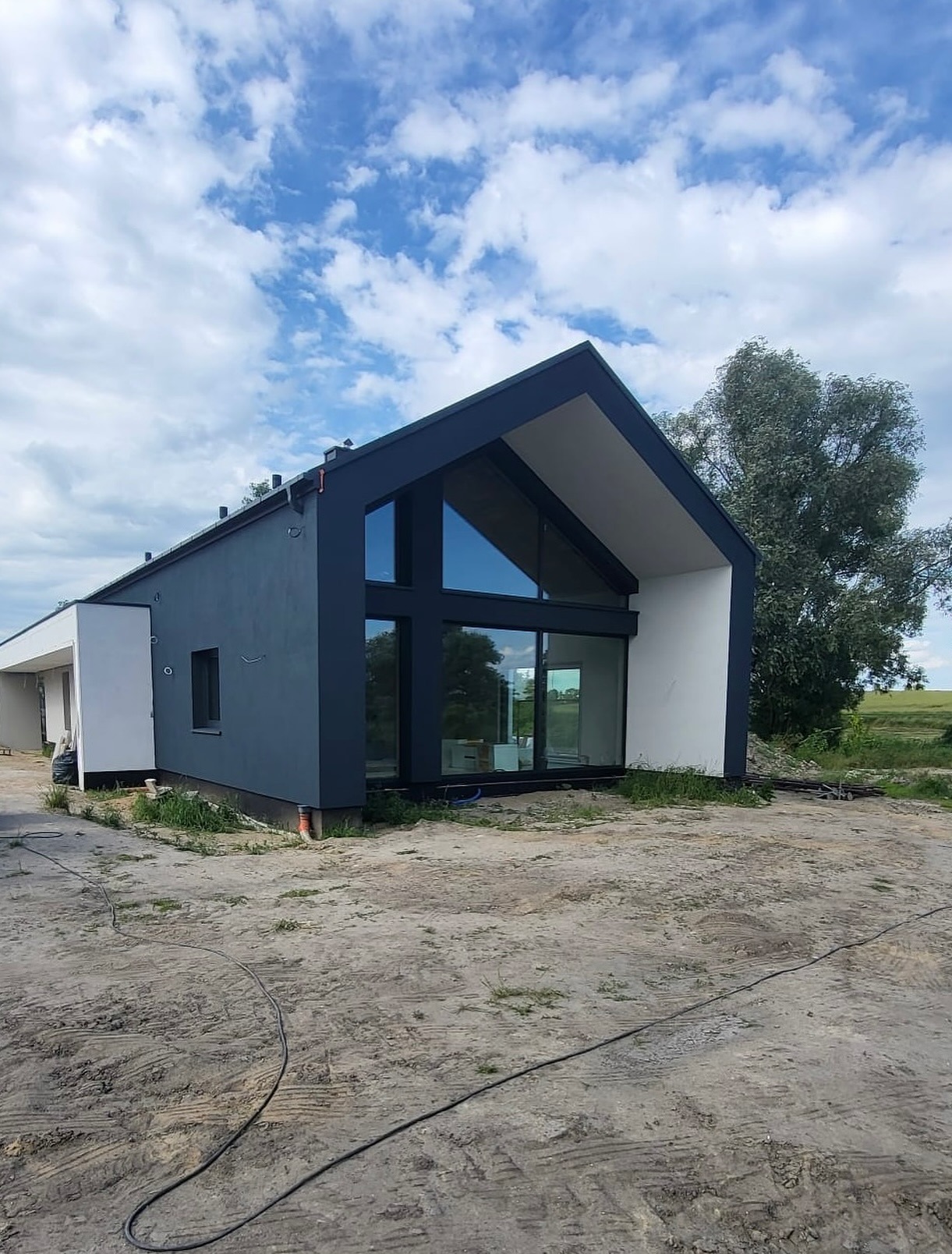 Nowoczesny dom jednorodzinny z ciemnoszarą elewacją i dużymi przeszkleniami, na tle błękitnego nieba z chmurami. Widok z zewnątrz, w otoczeniu zieleni.