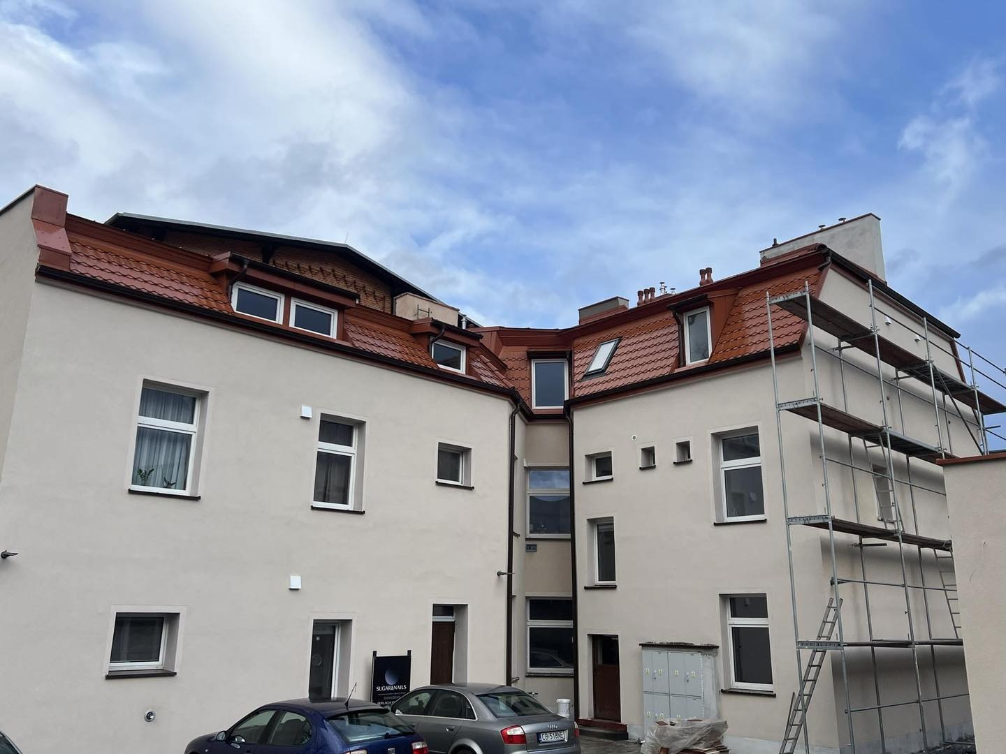 Budynek po renowacji dachu z nową, czerwoną dachówką, widoczne rusztowania i zaparkowane samochody.