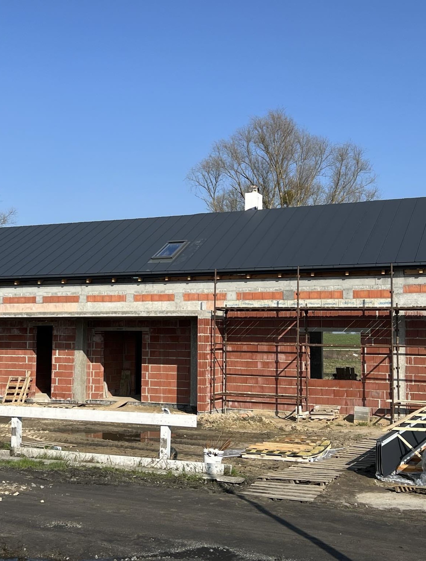 Dom w trakcie budowy z czarnym, ukończonym dachem z oknem dachowym, widoczne rusztowania i ceglana konstrukcja ścian.