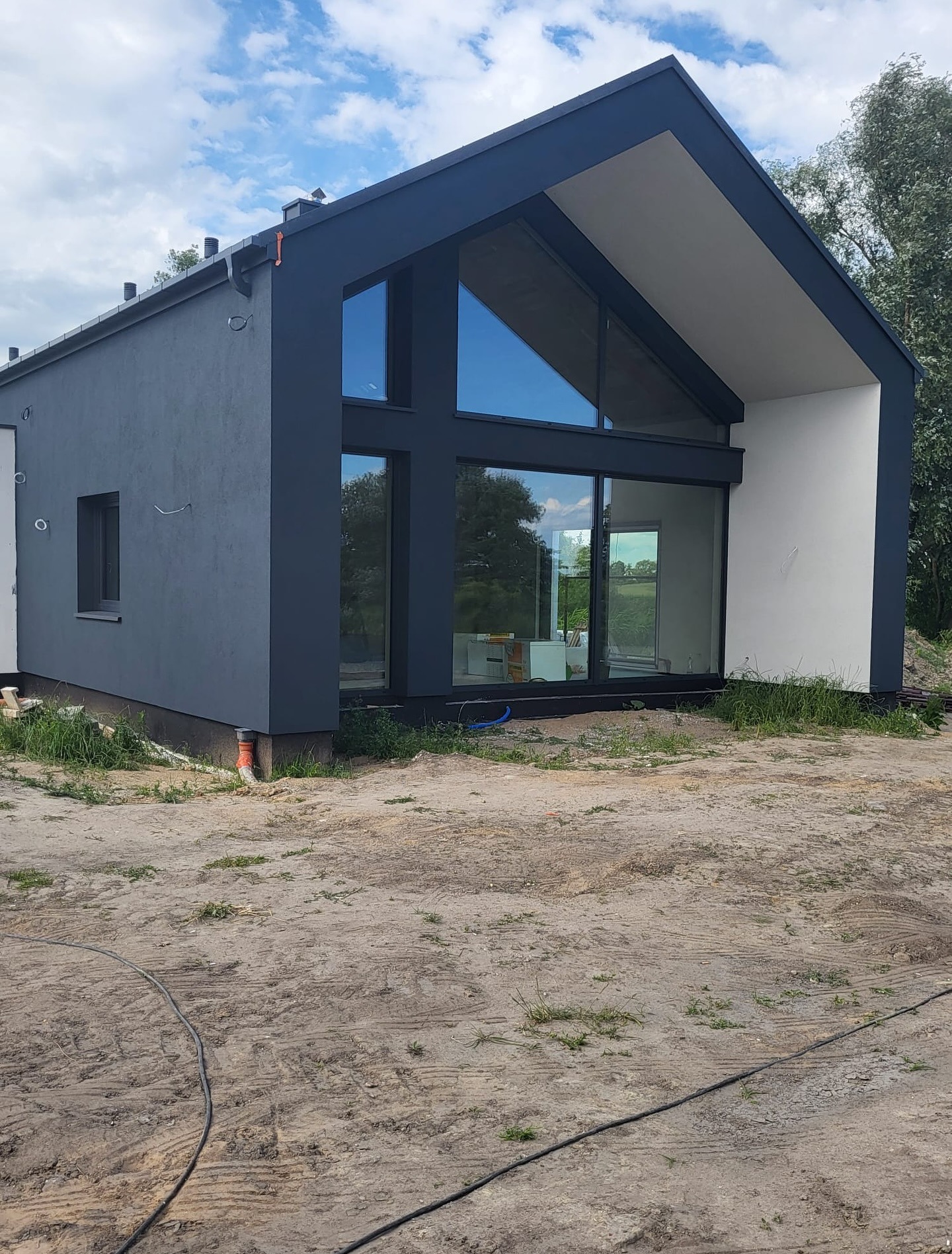 Nowoczesny dom z ciemnoszarą elewacją i dużymi oknami, widoczny fragment dachu i rynny, otoczenie w trakcie prac wykończeniowych.