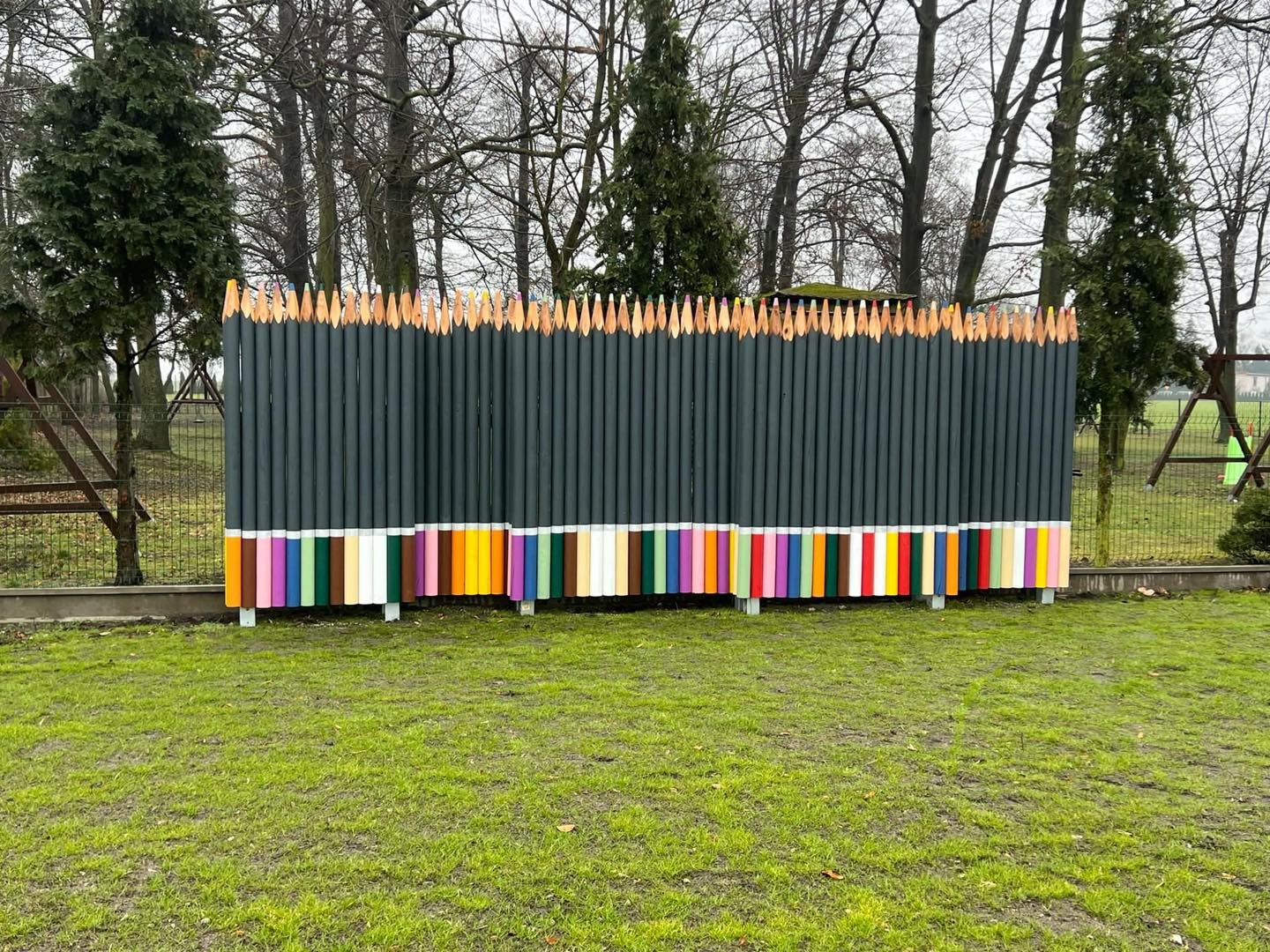 Oryginalne ogrodzenie z drewnianych kredek w różnych kolorach, otoczone zieloną trawą i drzewami bez liści w tle. Nietypowy design.