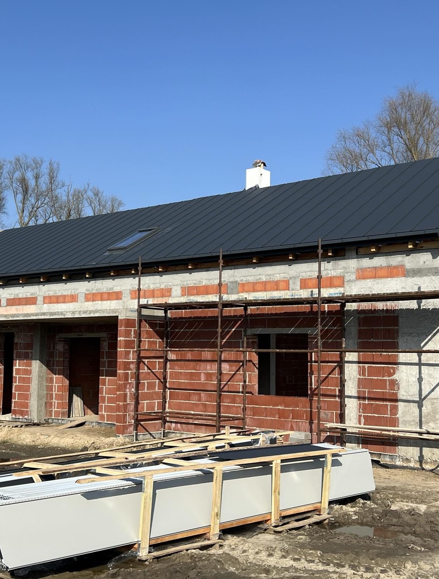 Budowa domu jednorodzinnego z ciemnym dachem z blachy na tle błękitnego nieba. Widoczne rusztowania i materiały budowlane.