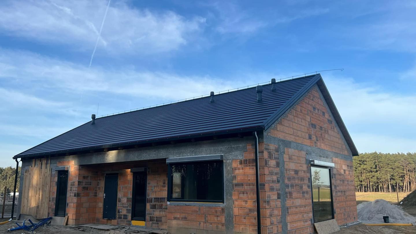 Dom w trakcie budowy z nowym, ciemnym dachem z dachówki ceramicznej, widoczne kominy wentylacyjne i surowe mury z cegły. Błękitne niebo w tle.