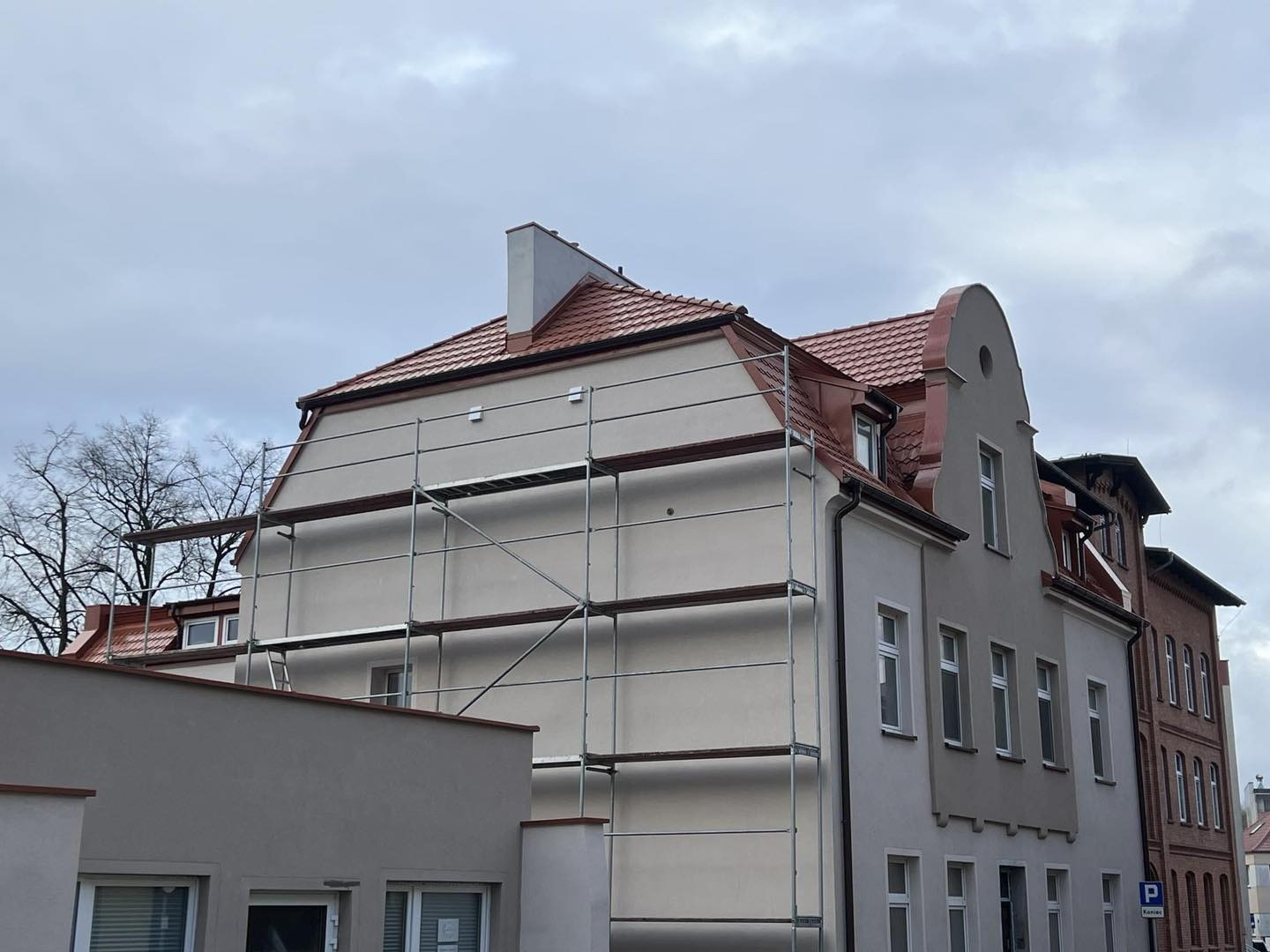 Budynek z nowym, czerwonym dachem w trakcie prac wykończeniowych. Widoczne rusztowania i fragmenty elewacji. Pochmurne niebo w tle.