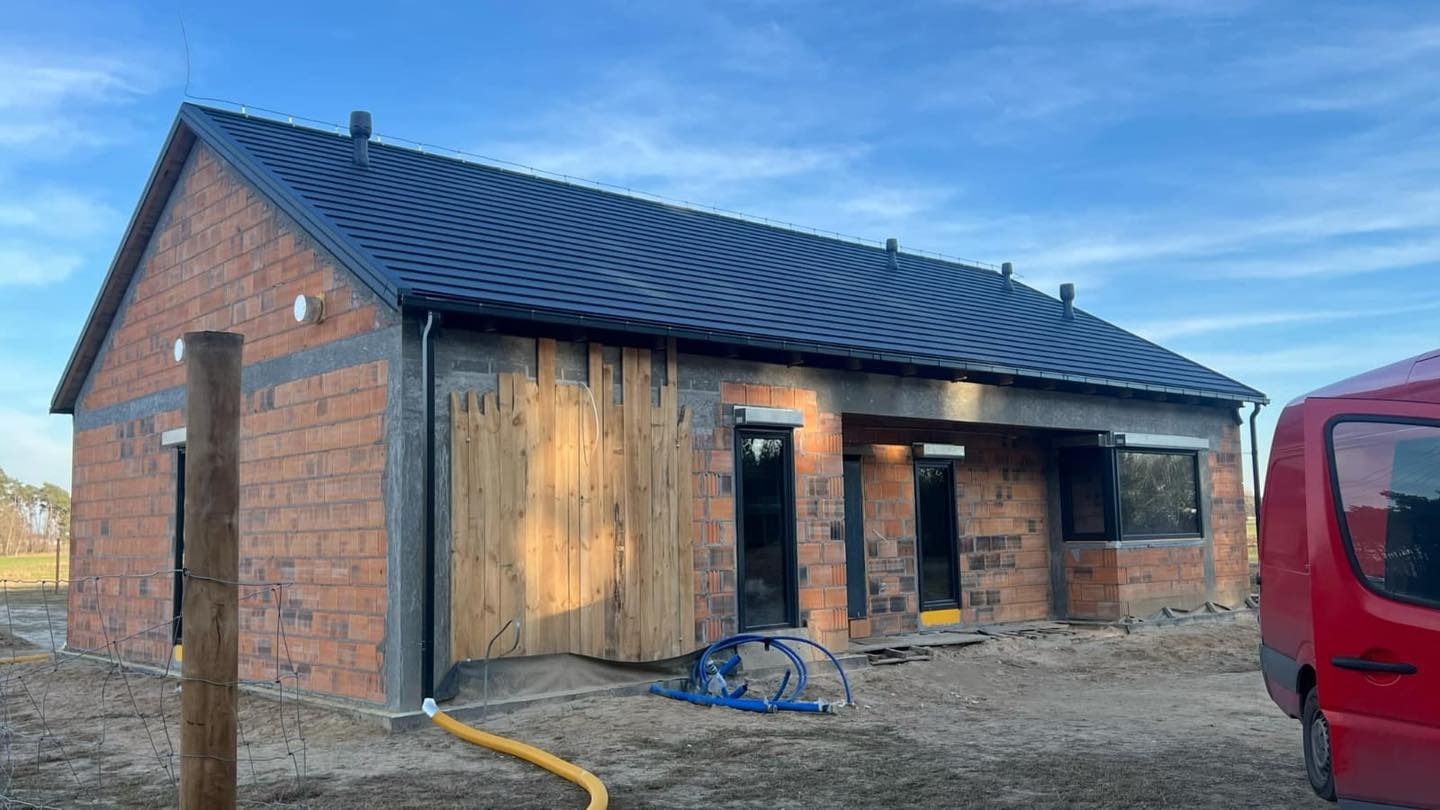 Dom jednorodzinny w trakcie budowy z ciemnym, nowym dachem. Widoczne detale: cegła, drewniane elementy, okna z roletami zewnętrznymi. Błękitne niebo w tle.